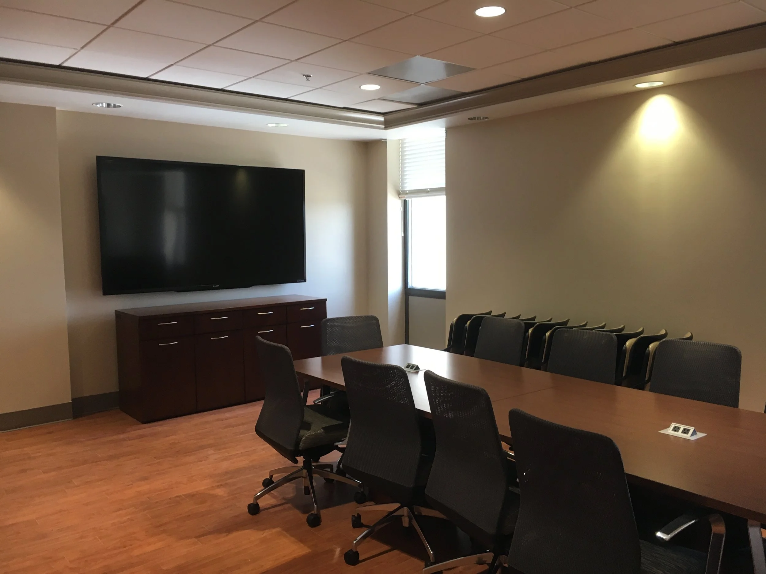 Conference room with a large wall-mounted flat-screen TV, a wooden conference table with several black ergonomic chairs, a window with blinds, and warm overhead lighting.