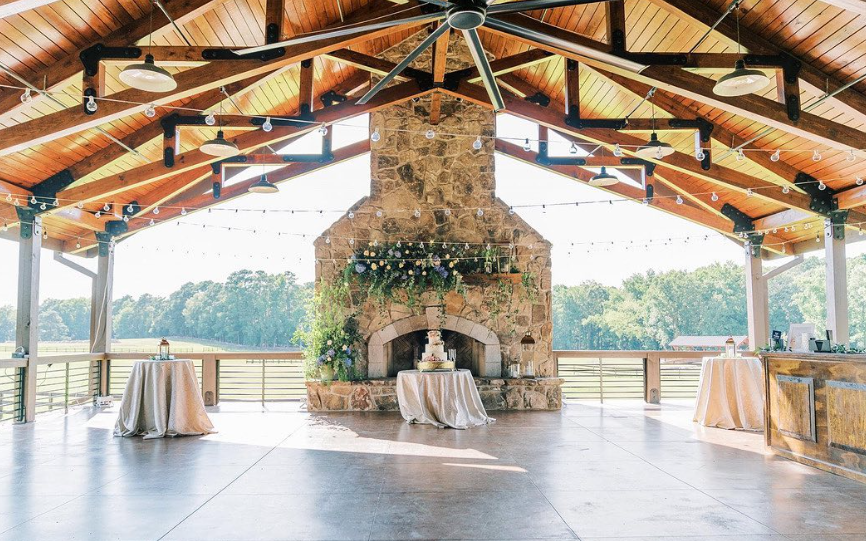 An open-air pavilion with a high wooden ceiling, stone fireplace decorated with greenery and string lights, and a cake on a table in front of the fireplace, set up for an event with tables covered in white cloths.