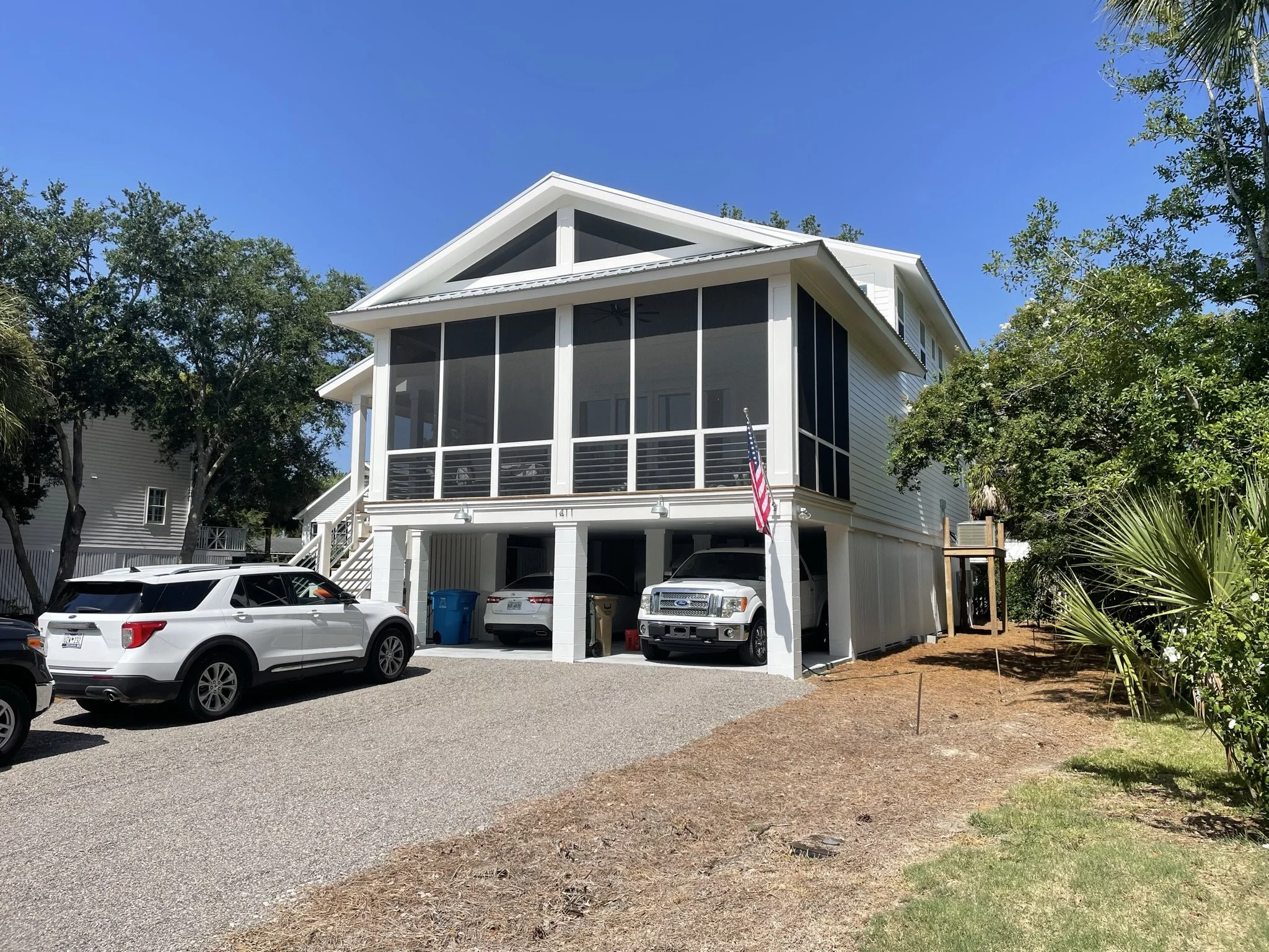 A white multi-story house with a screened-in porch, a garage at the bottom, a small American flag on a pole, parked cars, trees, and a blue sky.