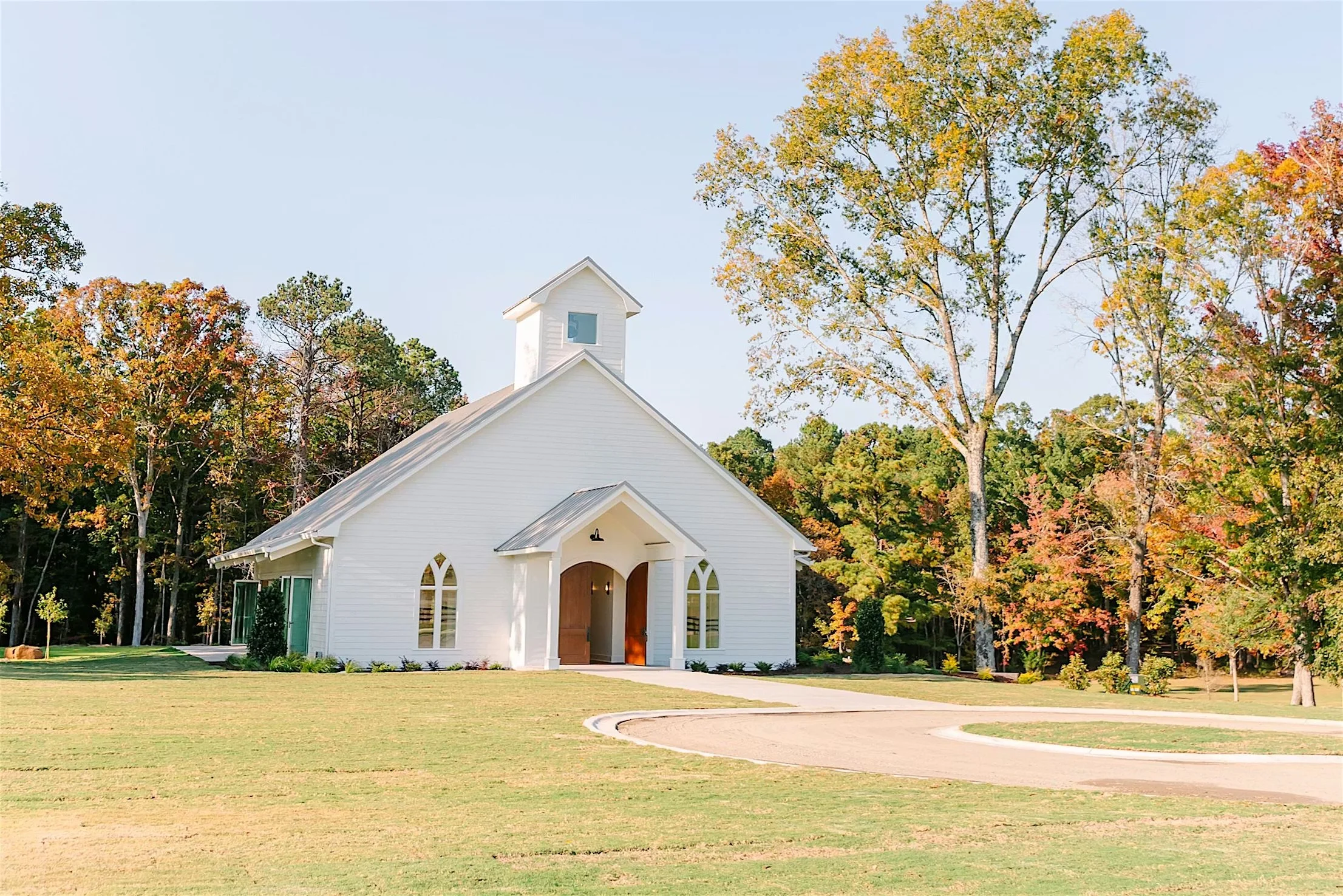 A small white chapel with a brown door, surrounded by trees with fall foliage, under a clear blue sky.