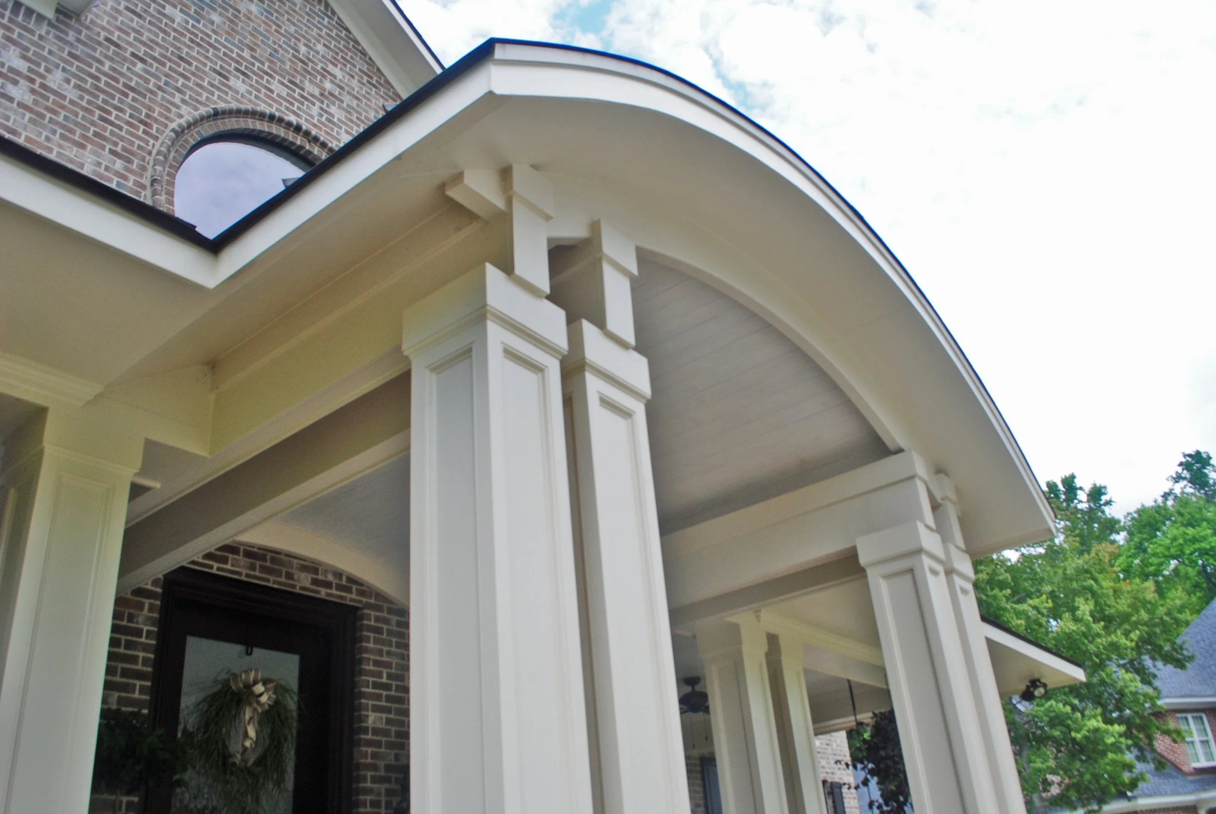 Close-up of a house porch with white columns and an arched roof, brick exterior wall, and a window, under a partly cloudy sky.