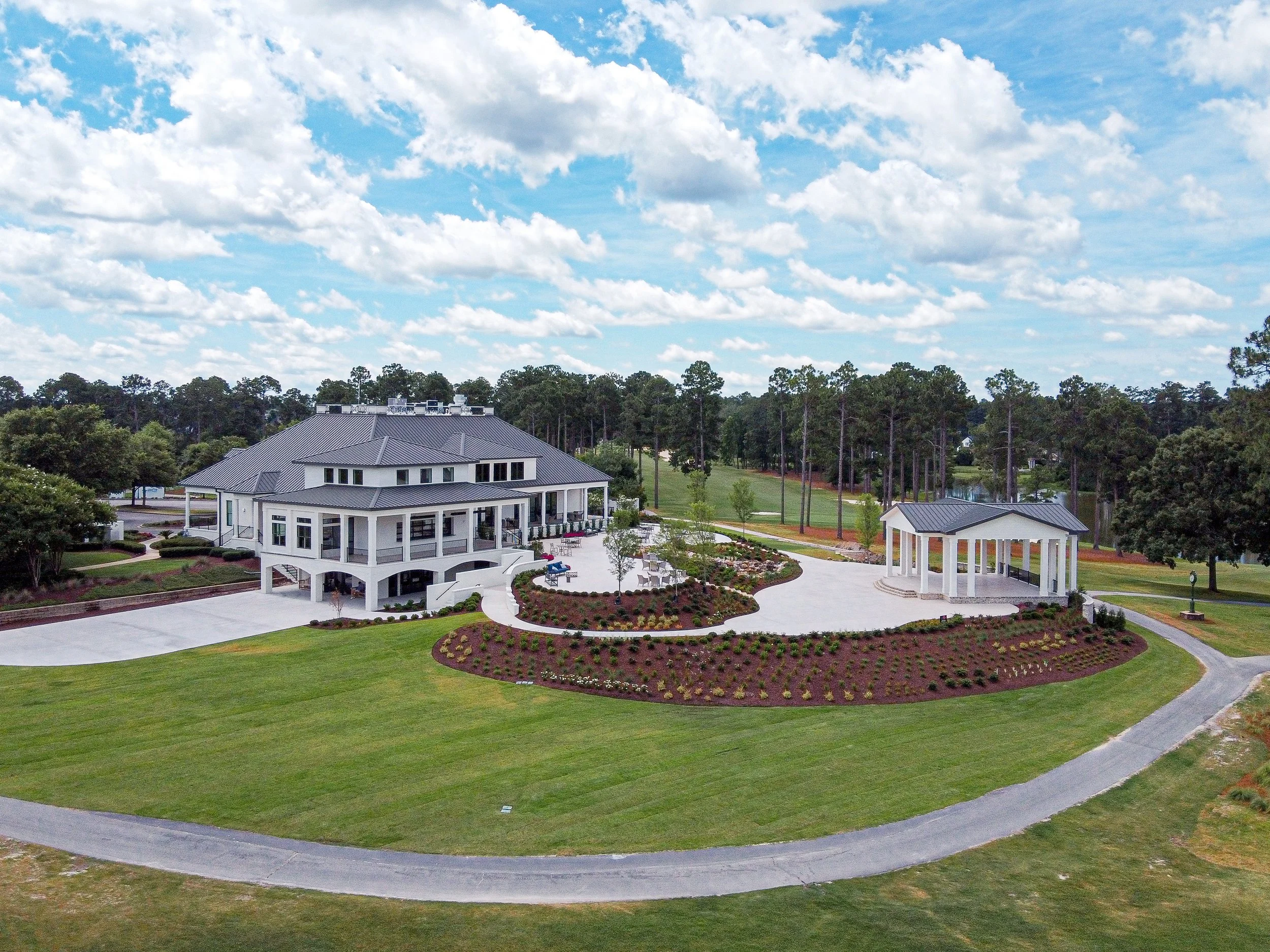A large house with a gray metal roof, white exterior, multiple balconies, surrounded by a landscaped yard with a circular driveway, flower beds, and a small white pavilion. There is a dense forest of trees in the background under a partly cloudy sky.