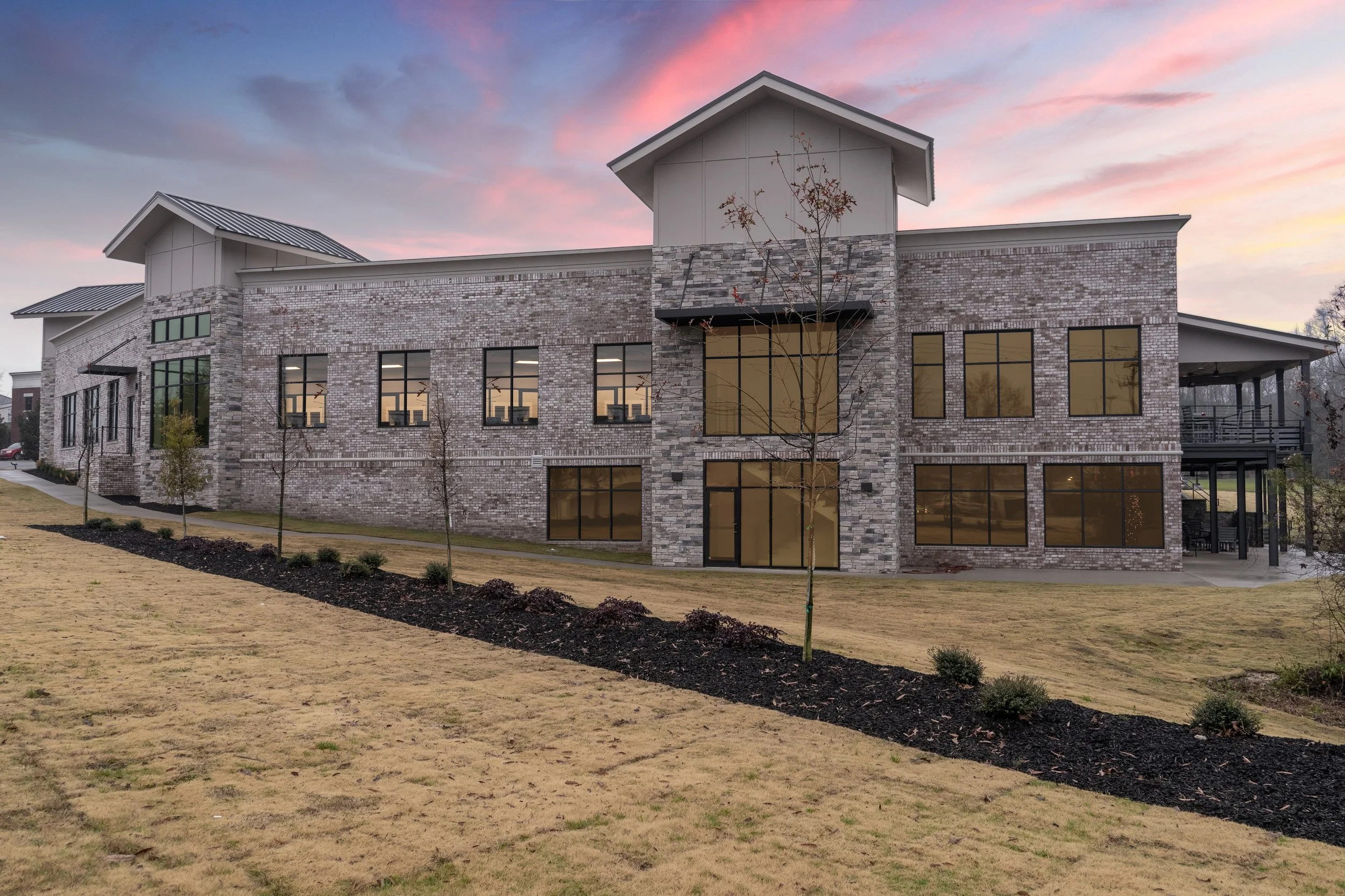 A modern multi-story building with brick exterior, large windows, and a sloped metal roof, set against a colorful sunset sky.