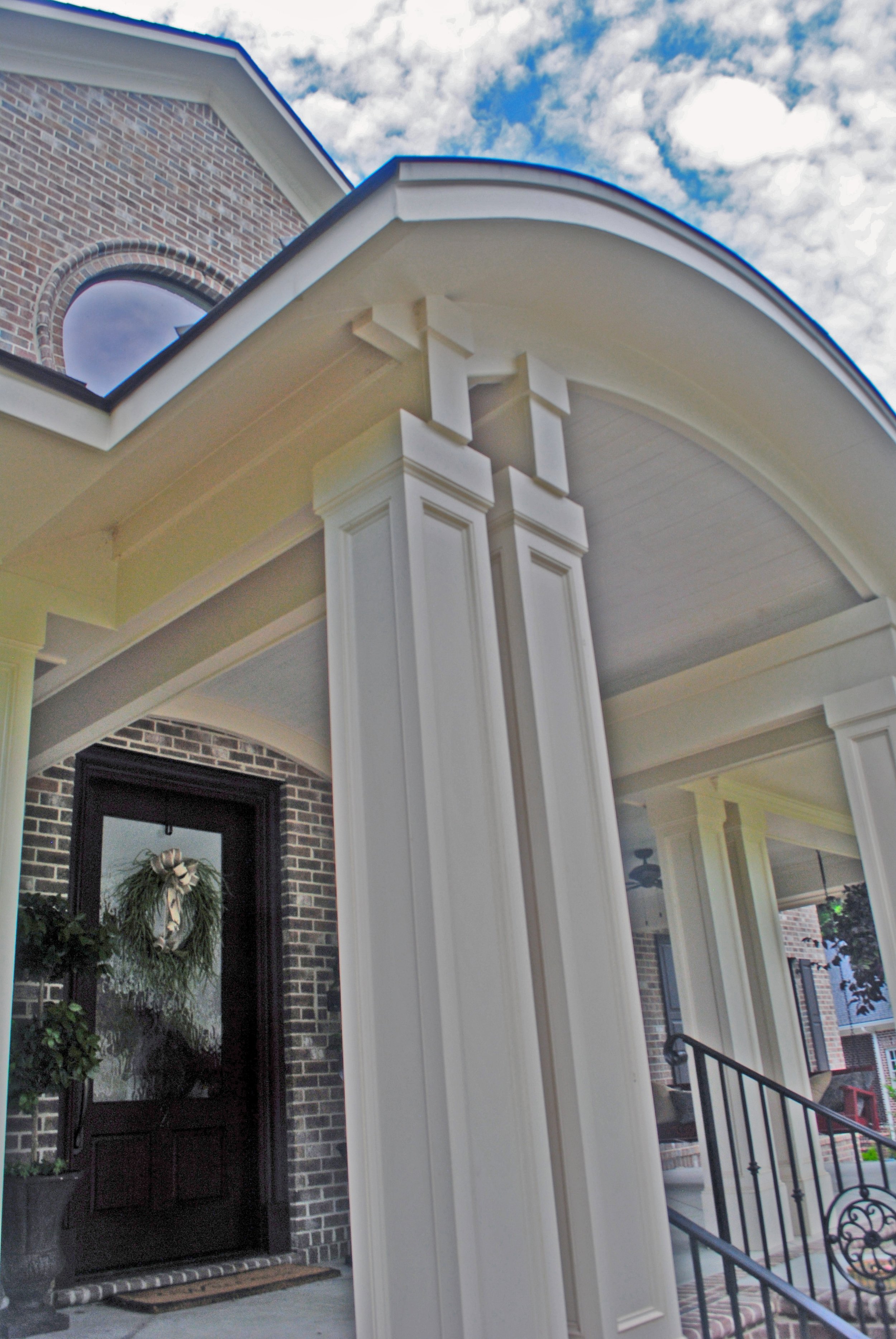 Close-up of brick house entrance with a black door decorated with a holiday wreath, and a porch with white columns and railing, under a partly cloudy sky.