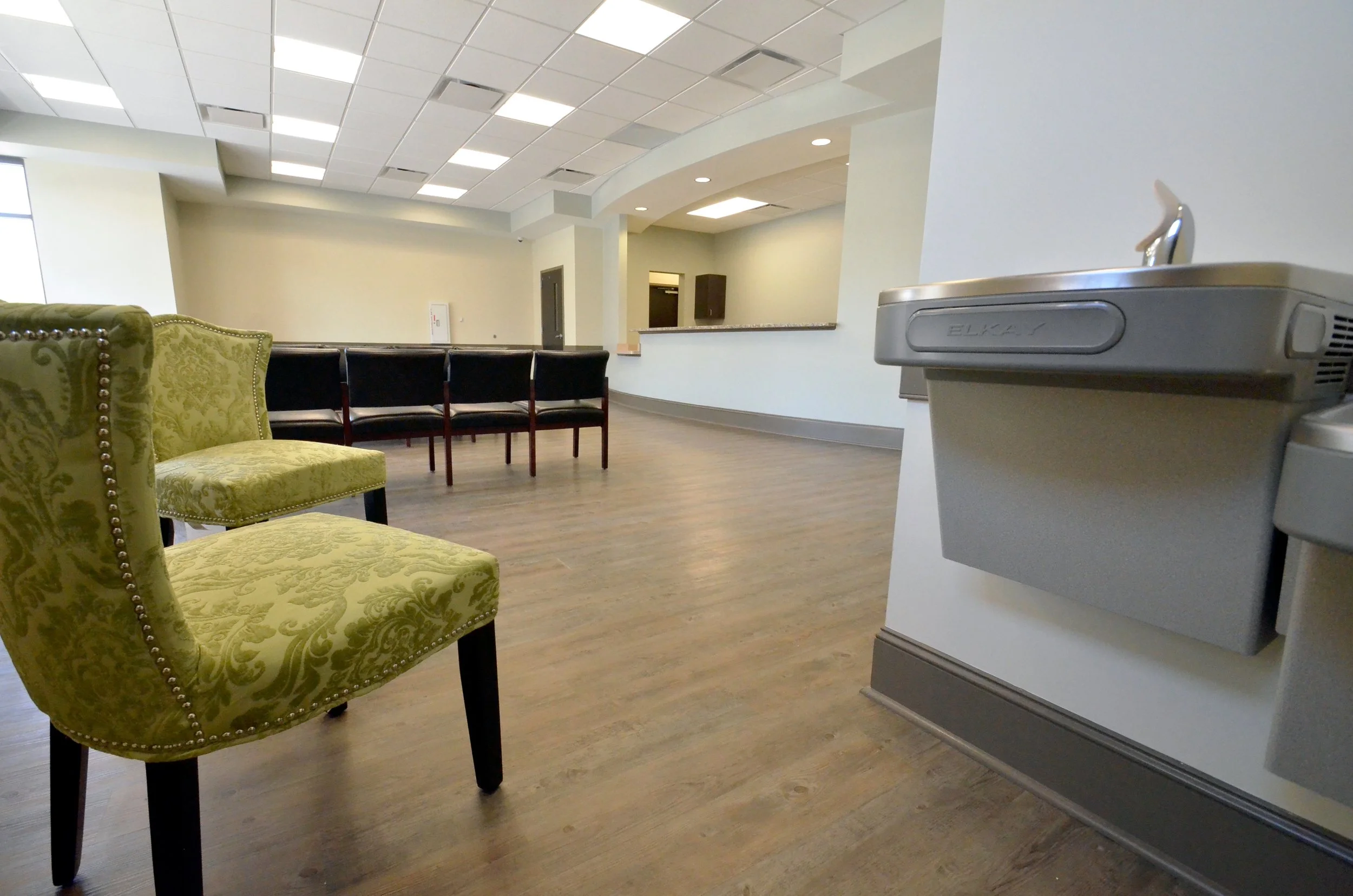 Empty waiting area with green upholstered armchairs, black chairs, and a white wall with a curved reception counter and a water fountain on the right.