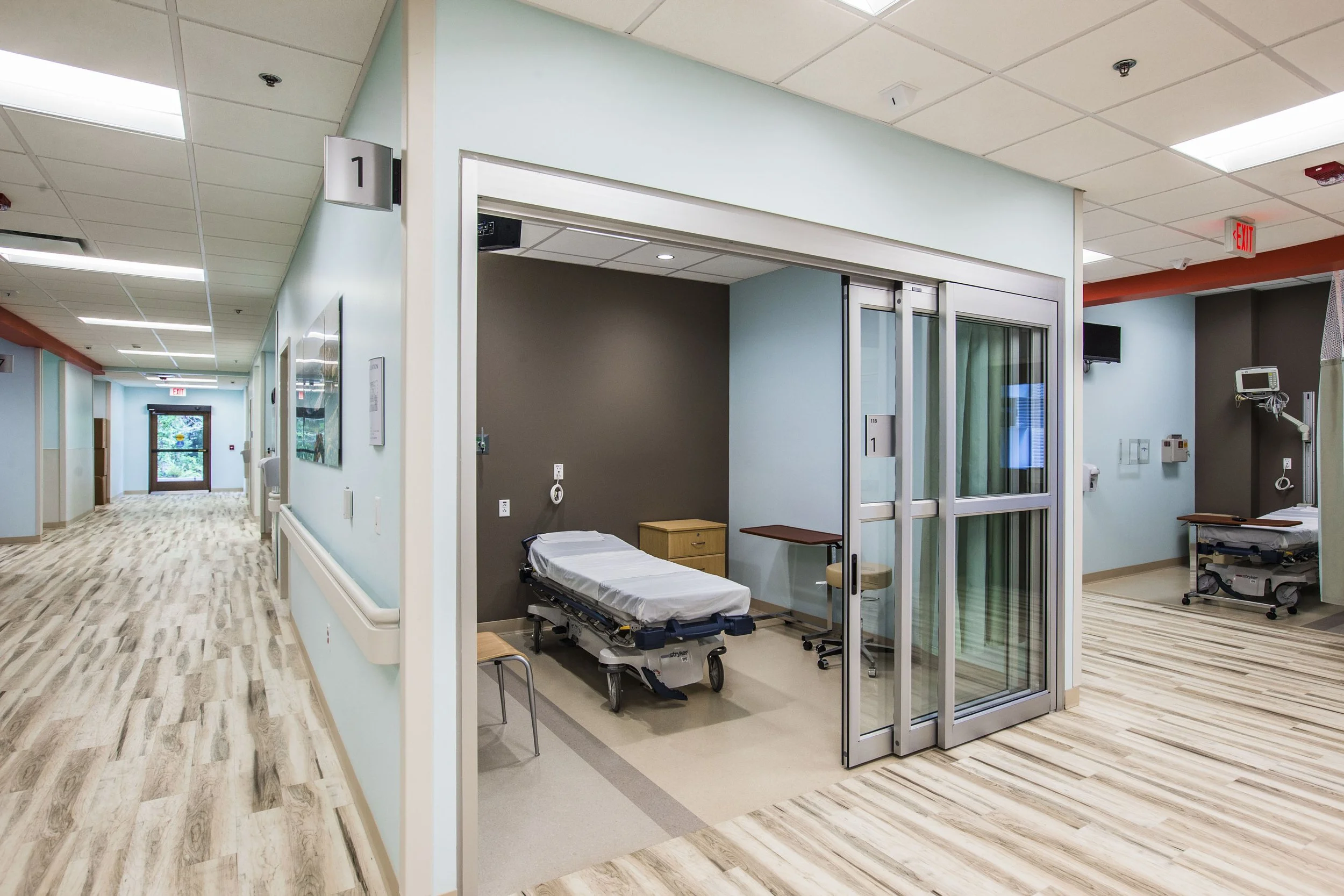 A hospital hallway with a patient room that has a sliding glass door, a hospital bed, medical equipment, and a TV mounted on the wall.