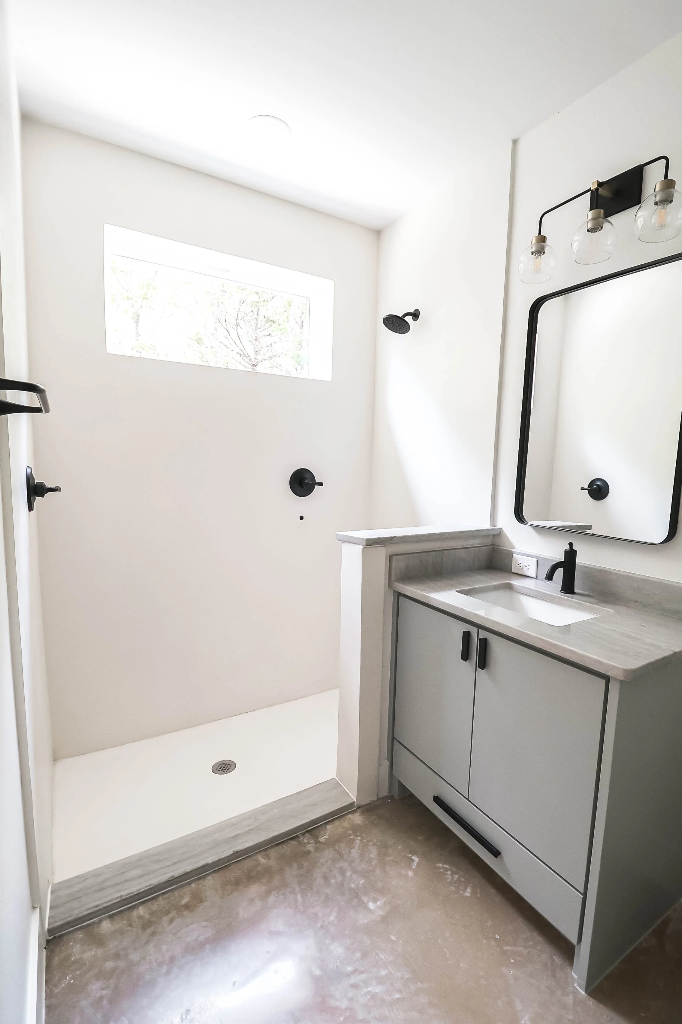 Modern bathroom featuring a white shower area with a black showerhead, a black-framed rectangular mirror, a gray vanity with black hardware, a small rectangular sink, and a wall-mounted light fixture with three glass shades. The wall is painted white