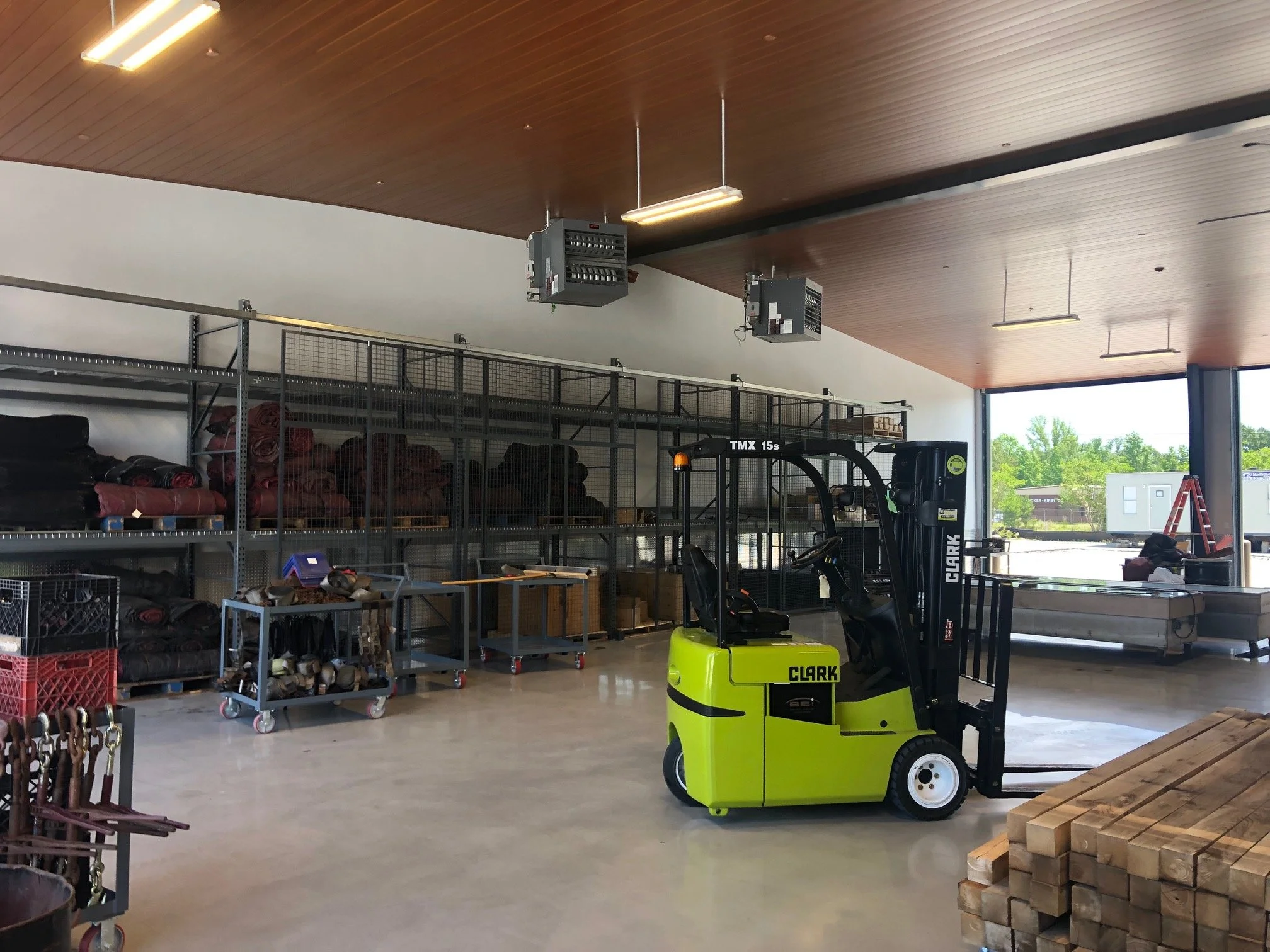 Interior view of a storage or warehouse area with a wooden ceiling, fluorescent lights, metal shelves, and various equipment including a green Clark forklift, stacks of bricks or blocks, and rolling carts.