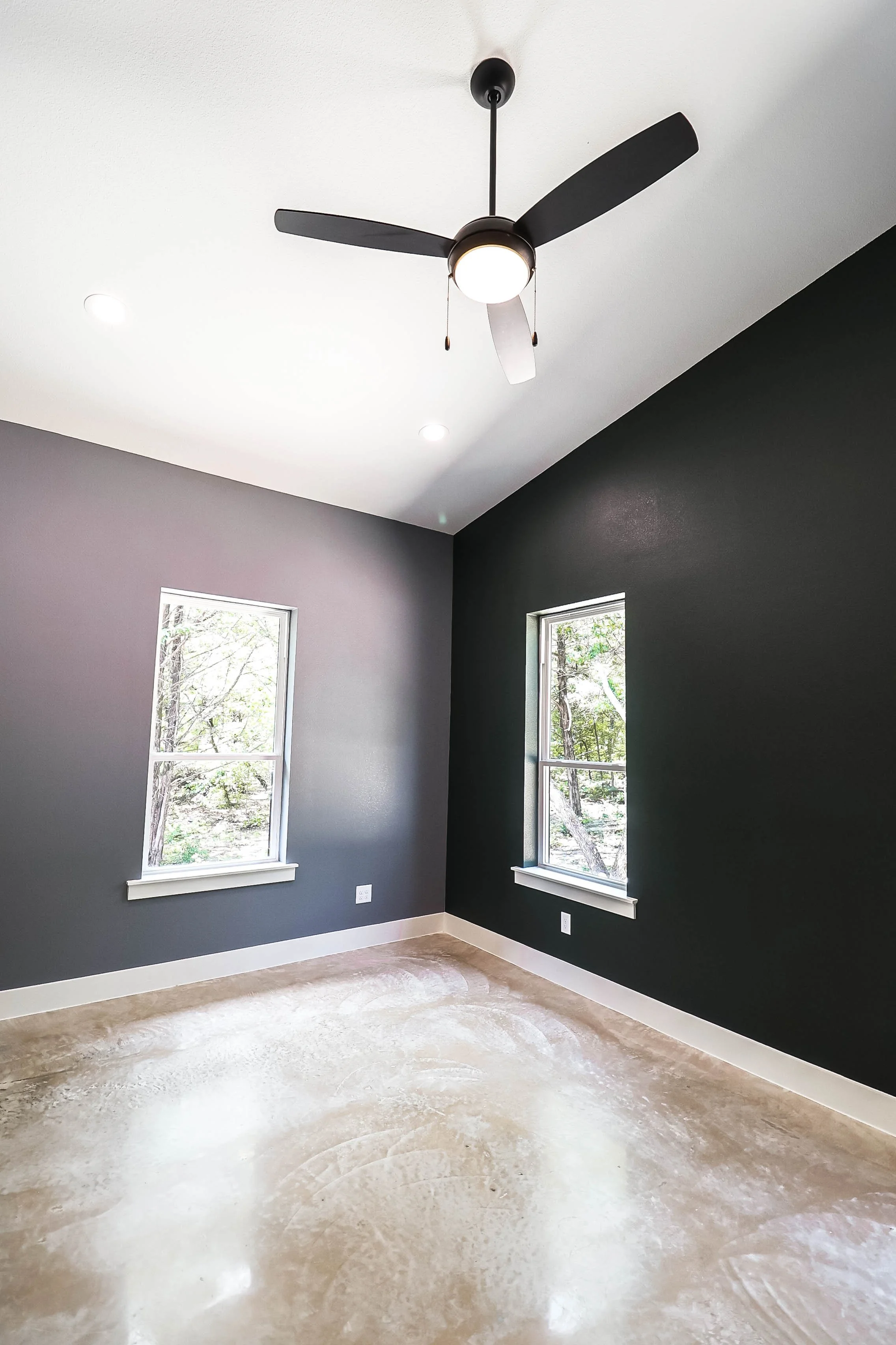 Empty room with two windows, dark gray and white walls, and a ceiling fan with a light. The floor is unfinished with a concrete surface.