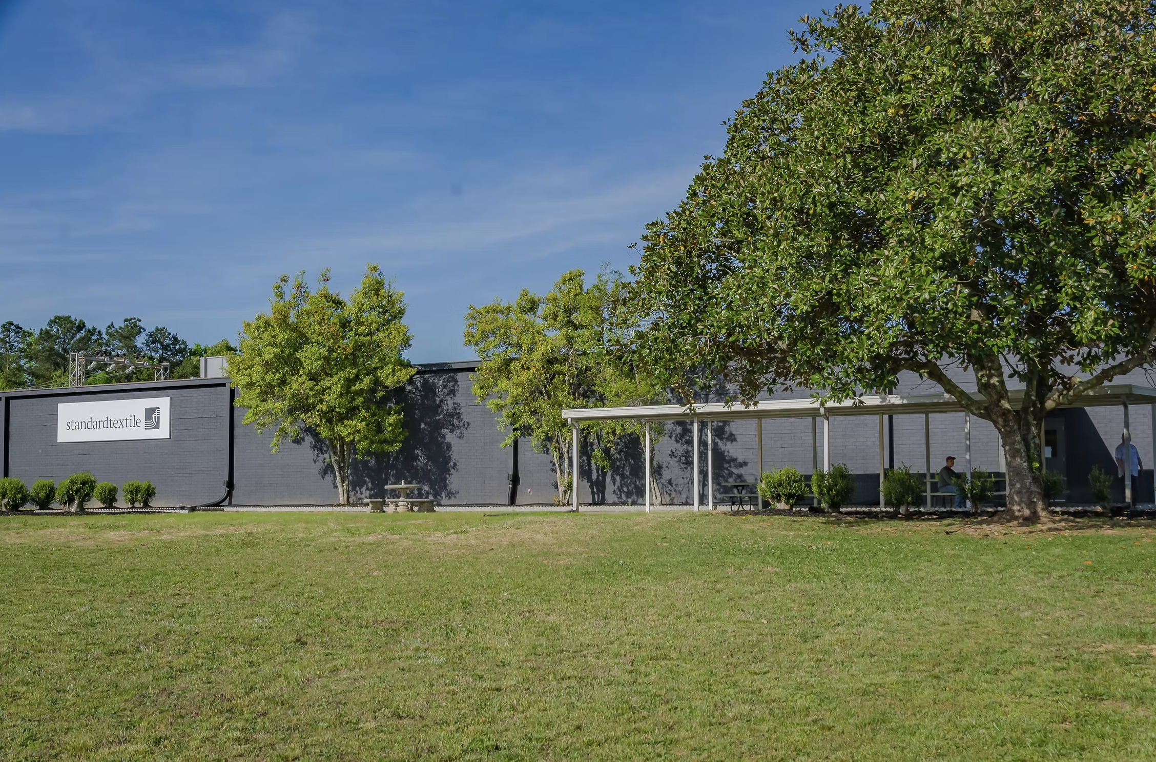 A modern gray building with a sign reading 'standard textile' and surrounded by a grassy lawn, five trees, and a clear blue sky.