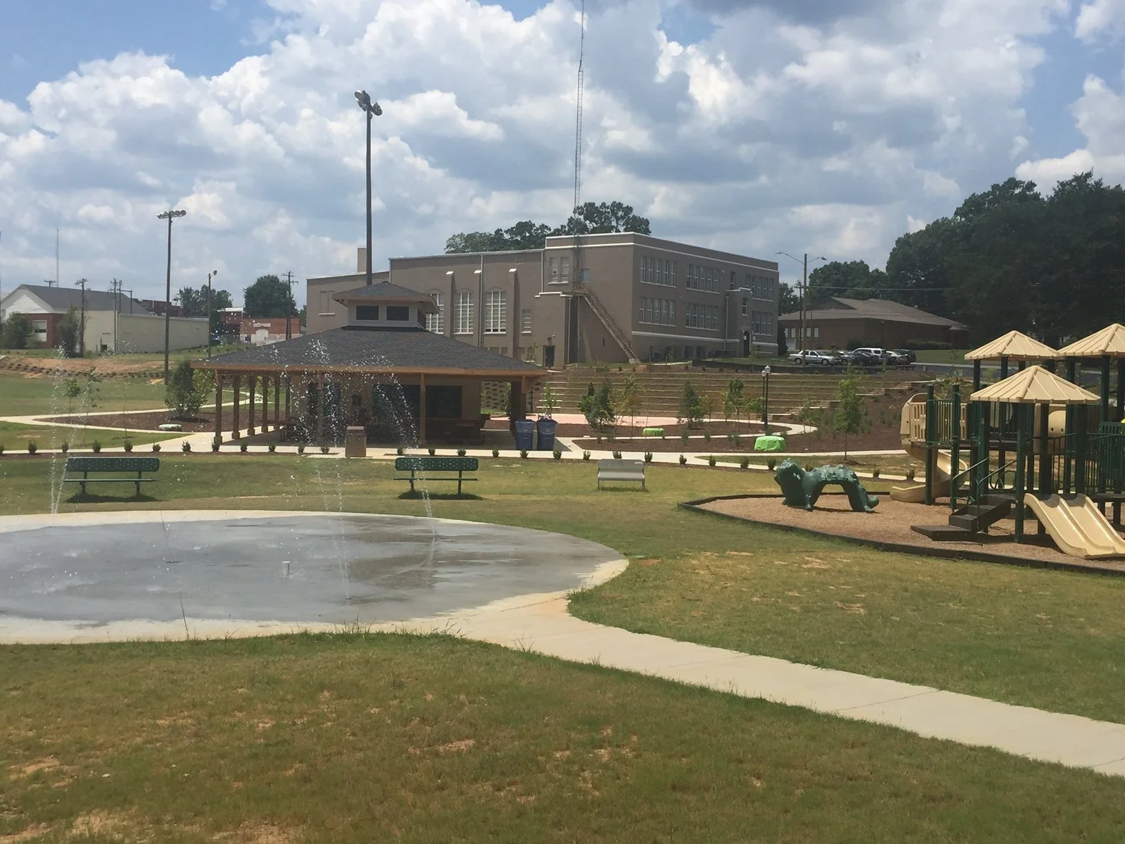 A park with a water fountain, benches, a playground, trees, and a building in the background under a cloudy sky.