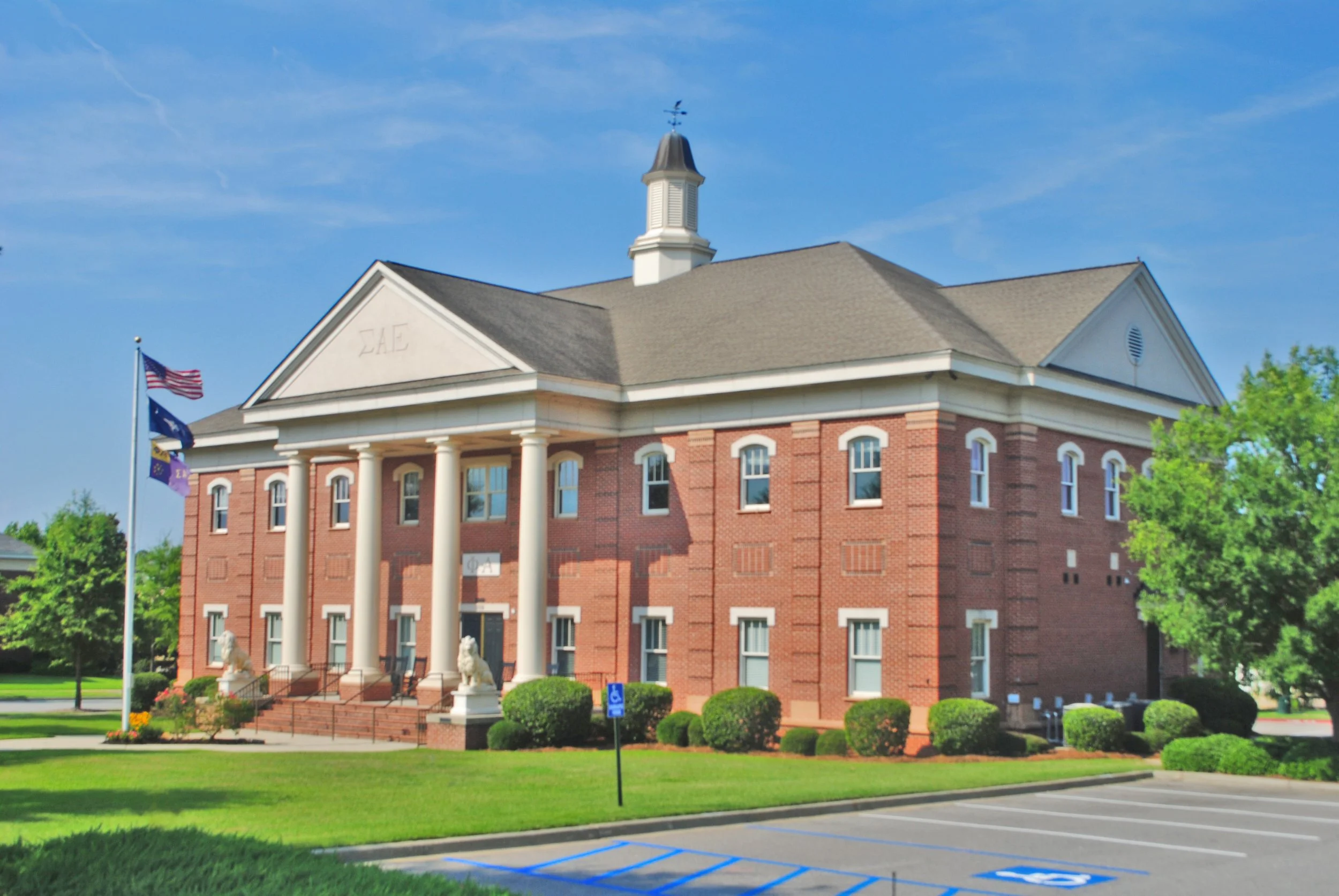 A red brick building with white columns and a statue of a lion in front. Flagpoles with American flags and a blue flag are nearby. The building has a steeple against a blue sky.