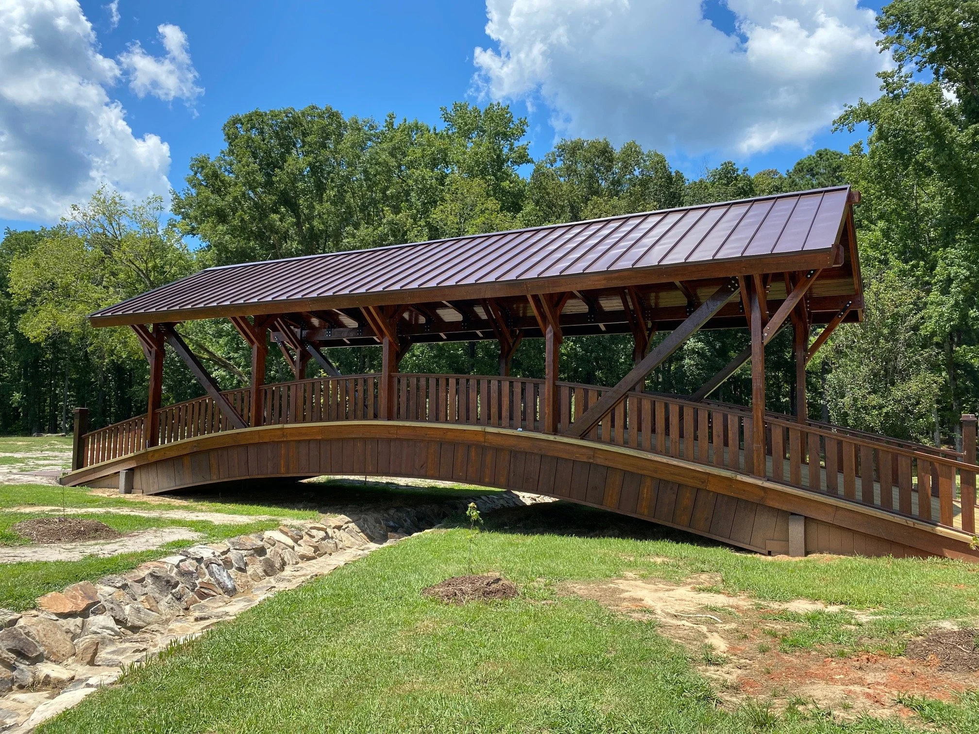 A wooden bridge with a curved deck and a sloped metal roof, surrounded by green trees and grass on a sunny day with partly cloudy blue sky.
