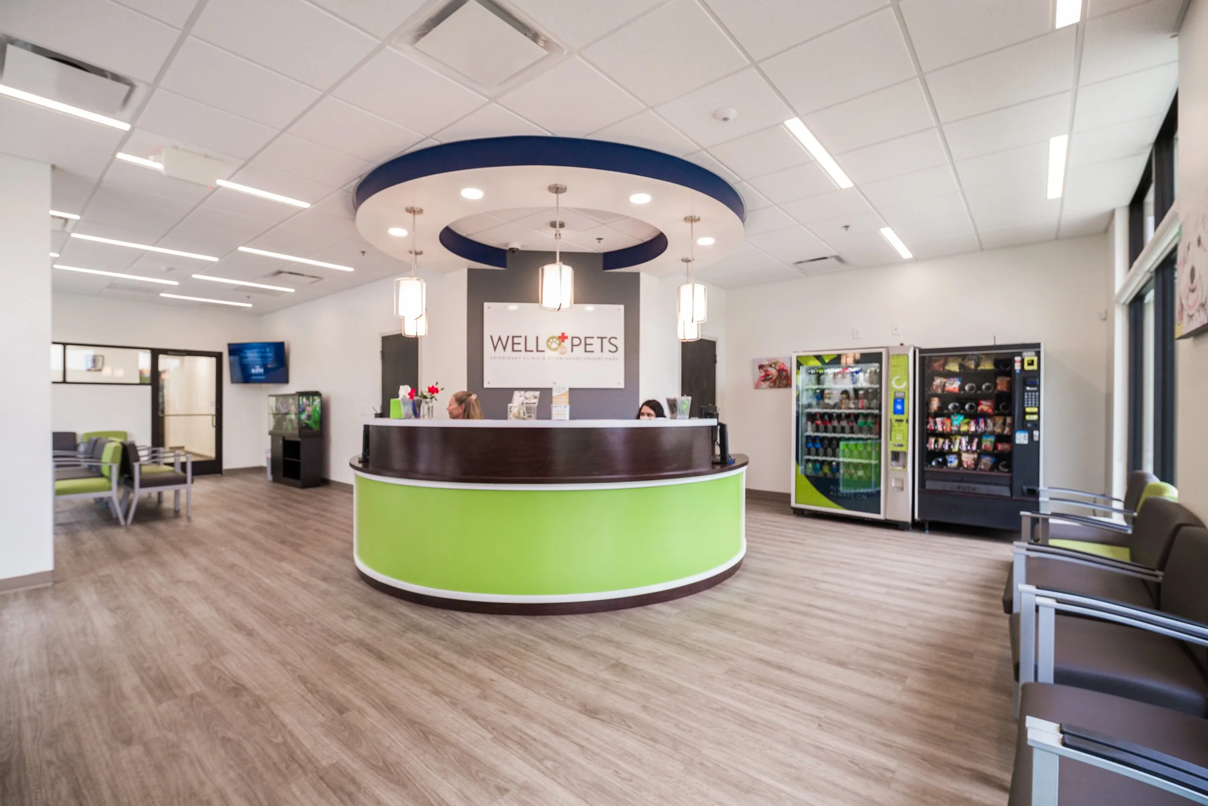 Reception area of Pet Wellness Center with modern decor, including a black and green curved reception desk, vending machines, seating area, and a wall-mounted TV, with a company sign reading "WELL PETS".