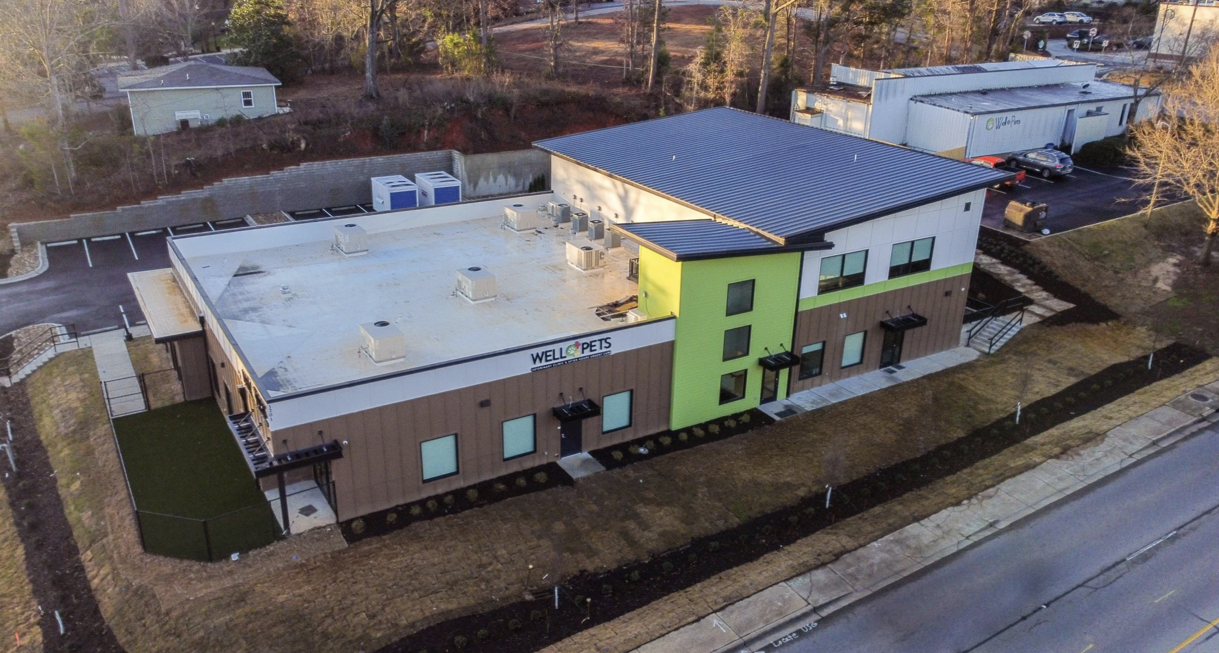 Aerial view of a modern two-story pet clinic building with the sign 'WELL PETS' on the side, surrounded by parking spaces, a sidewalk, and some landscaped areas with grass and young trees.
