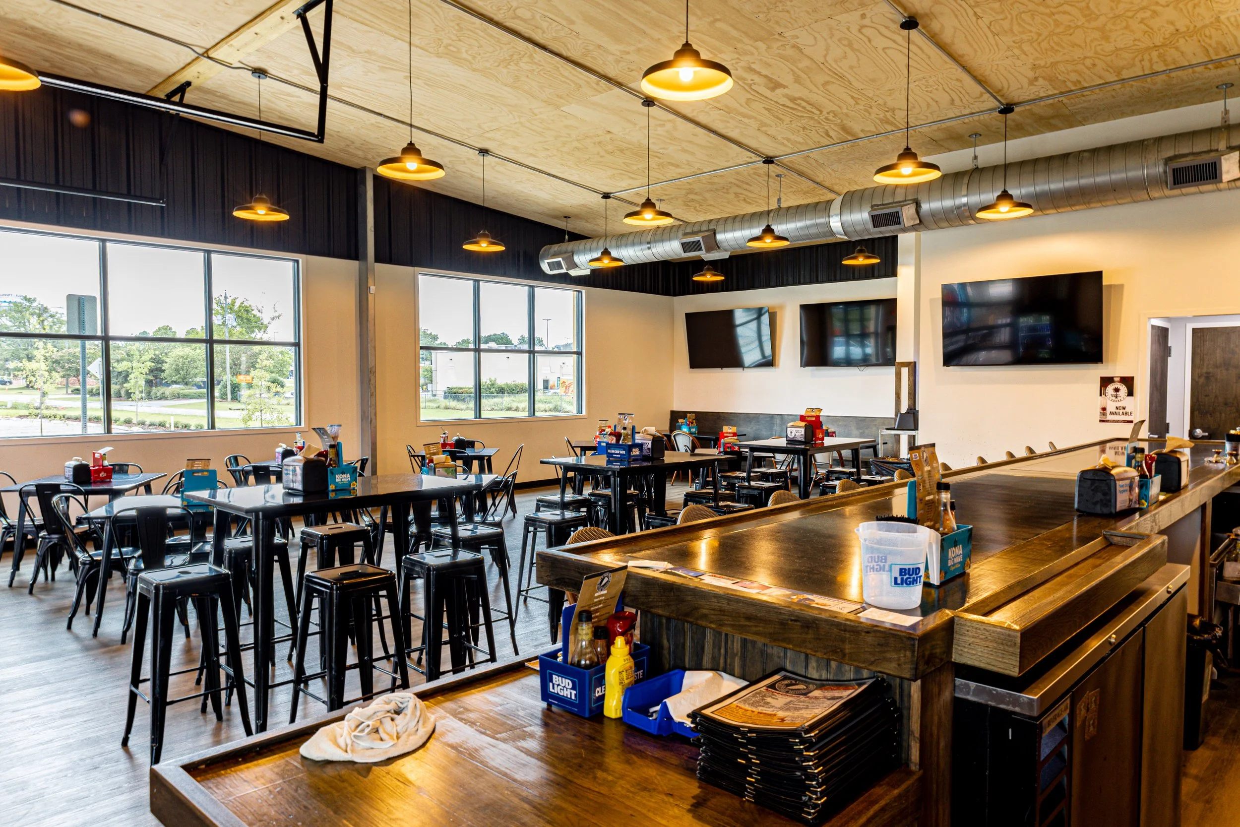 Interior of a restaurant or bar with high tables, chairs, condiment bottles, and two large flat-screen TVs on the wall. Large windows let in natural light, and decorative lighting fixtures hang from the wooden ceiling.
