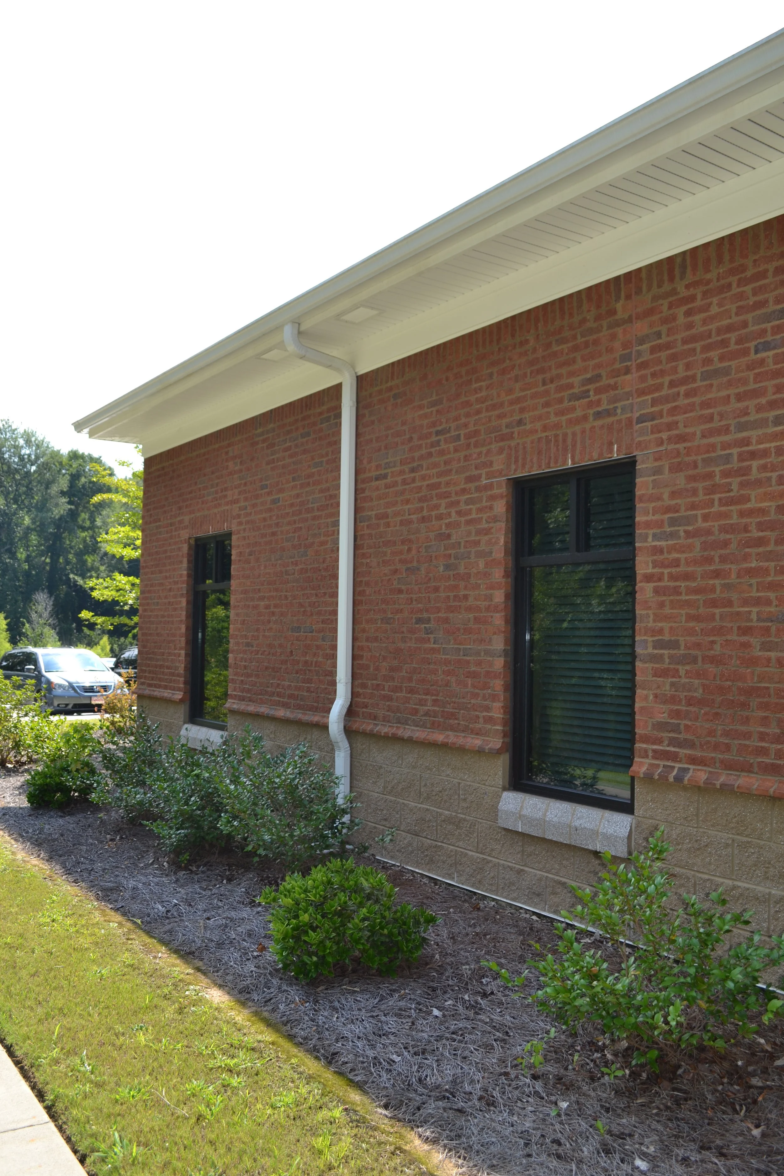 The side of a brick house with two black windows and a white gutter, surrounded by a landscaped yard with small shrubs and a grassy lawn.
