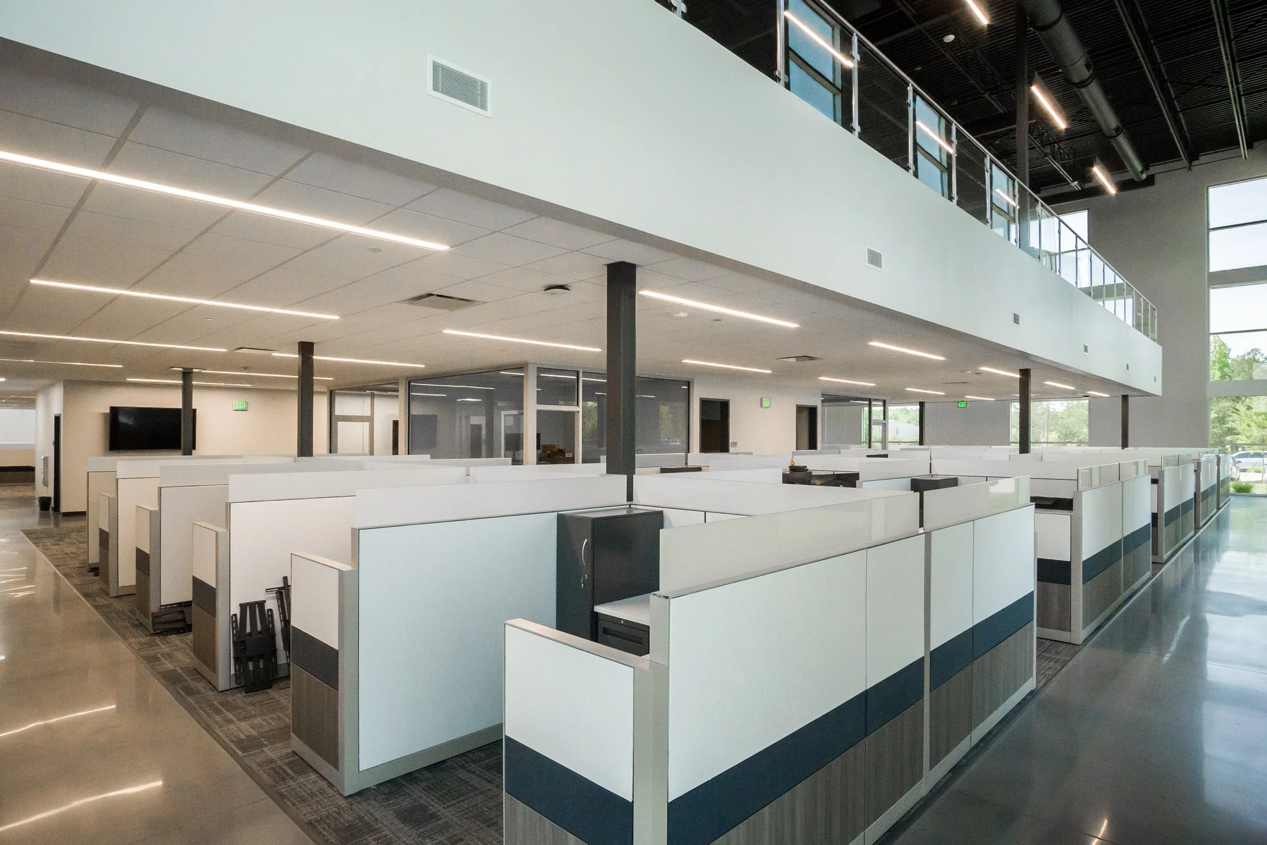 Open office space with rows of cubicles, a mezzanine walkway with glass-paneled railing, ceiling-mounted linear LED lights, and large windows showing trees outside.