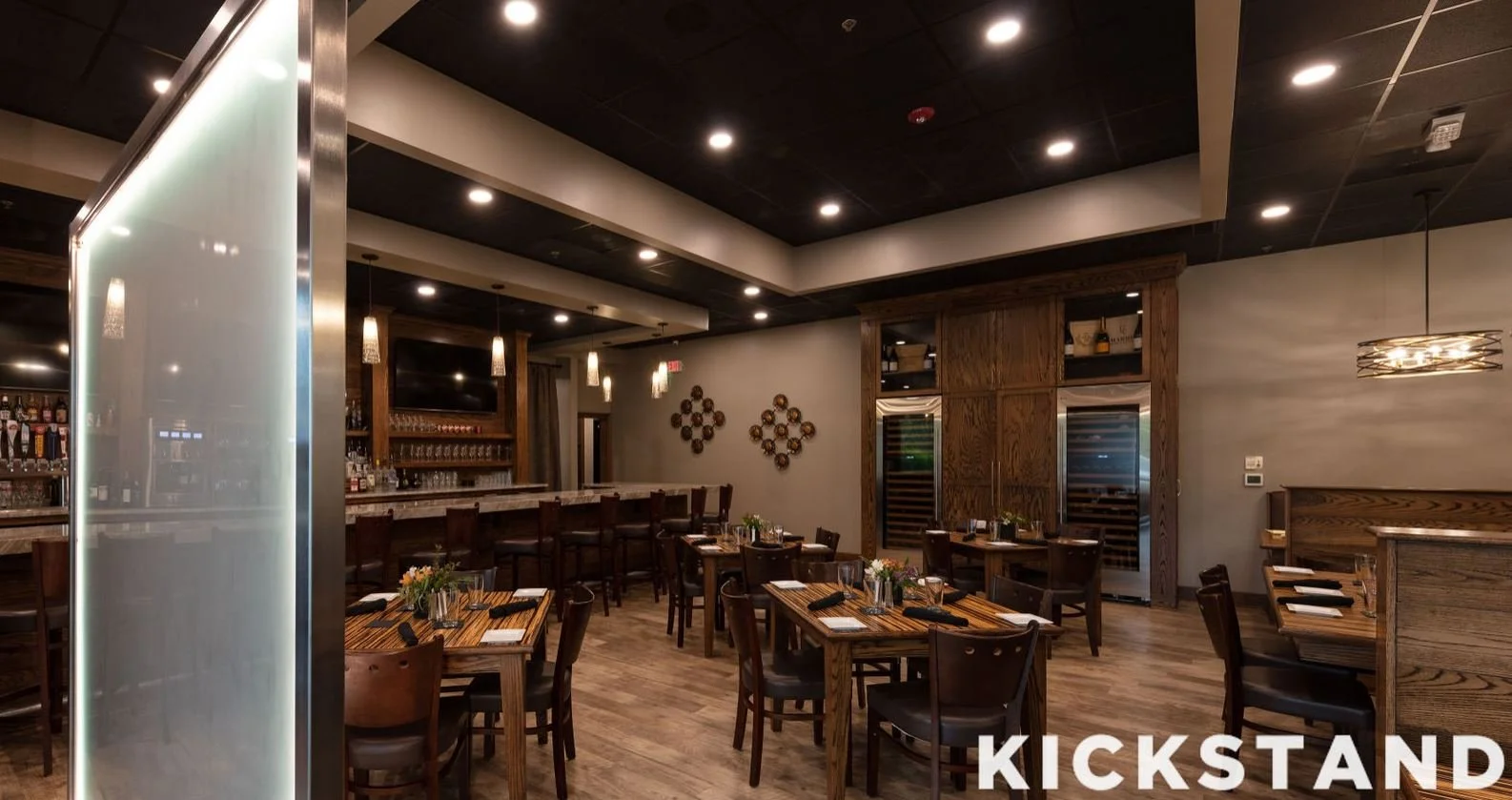 Interior of a modern restaurant with wooden tables and chairs, decorated with small flowers. Bar area with liquor bottles and wine coolers, warm lighting, and wall decorations.
