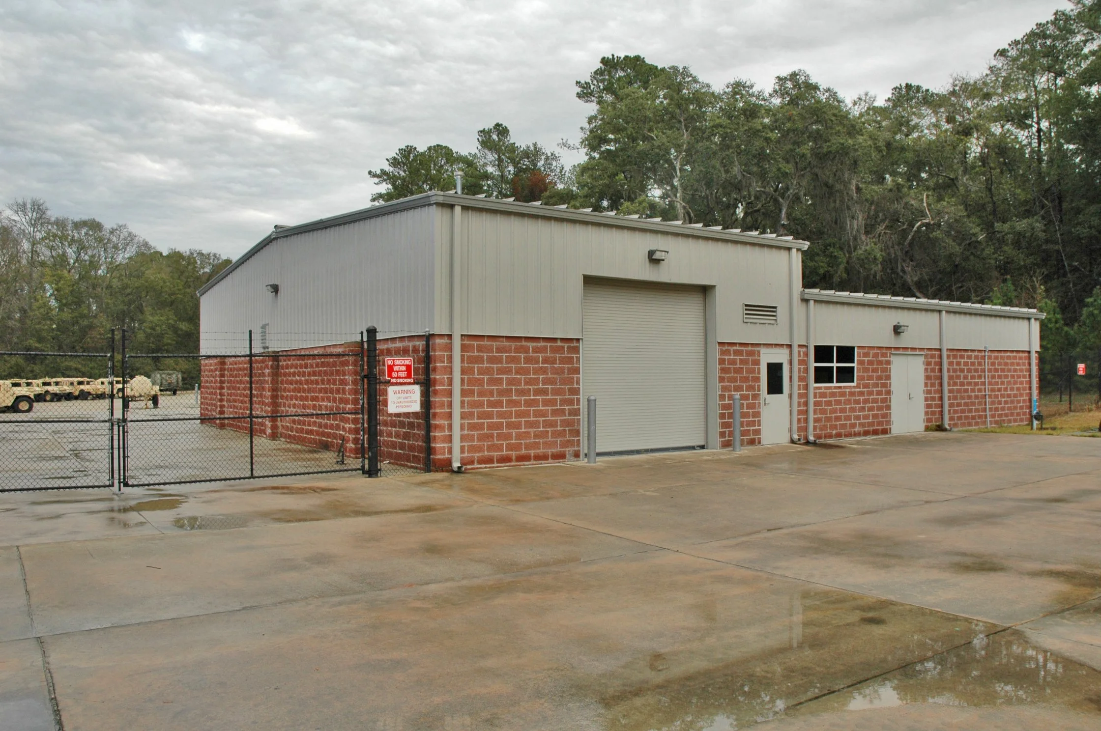 A large industrial metal building with a brick foundation, surrounded by a black chain-link fence, located in a parking lot with puddles. There are trees in the background under an overcast sky.