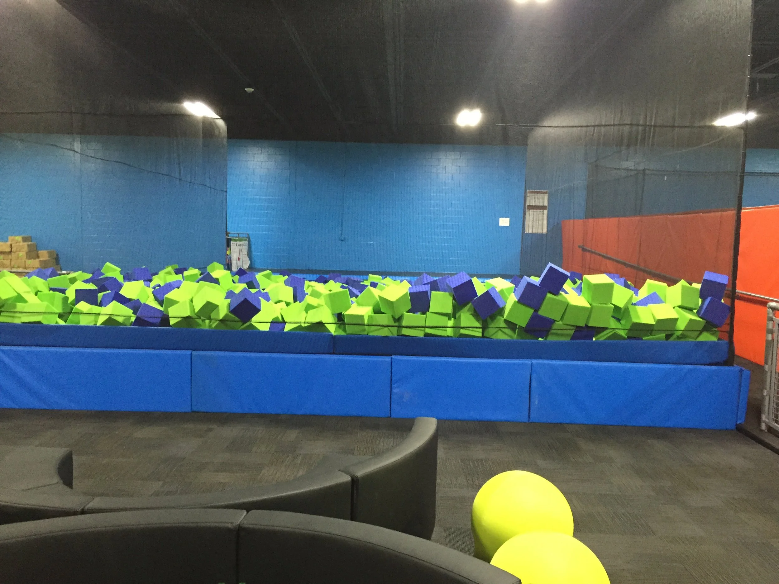Indoor trampoline park with foam pit filled with green and purple foam blocks and a yellow ball in the foreground.