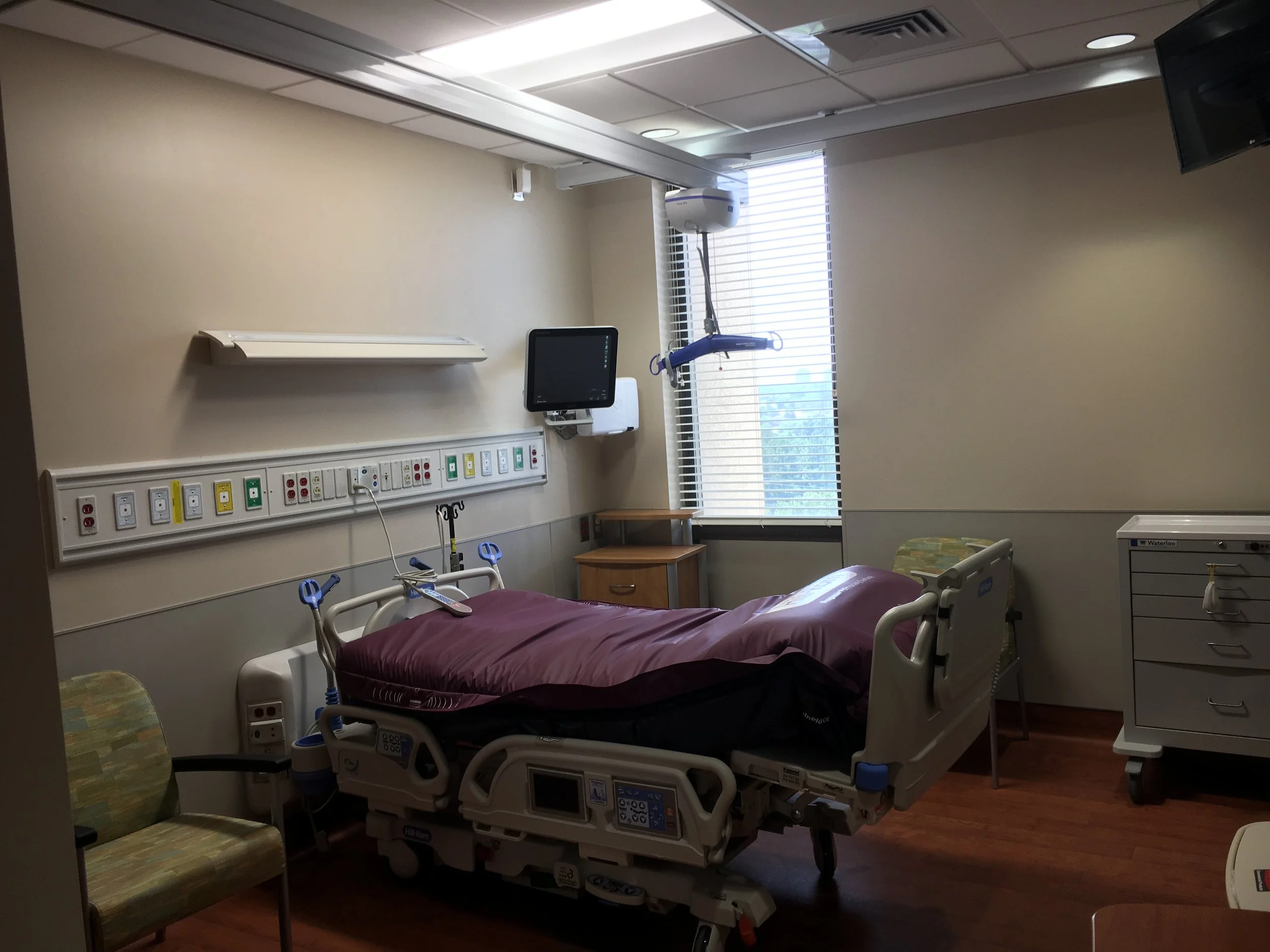 Hospital room with a bed, medical equipment, a window with closed blinds, and a chair.