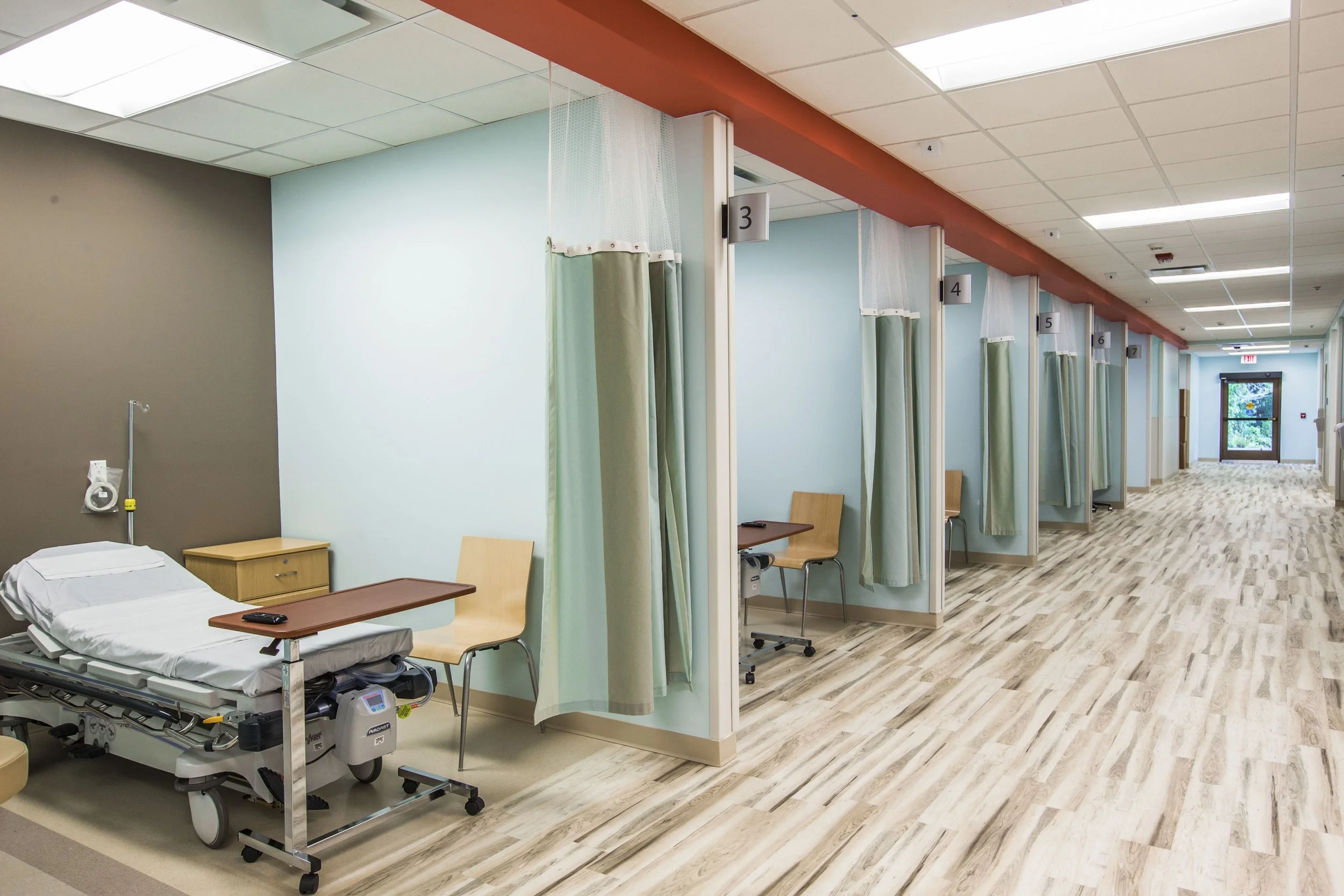 Hospital room with multiple patient beds separated by privacy curtains, numbered signs above each bed, wooden chairs, and a door with window at the end of the hallway.