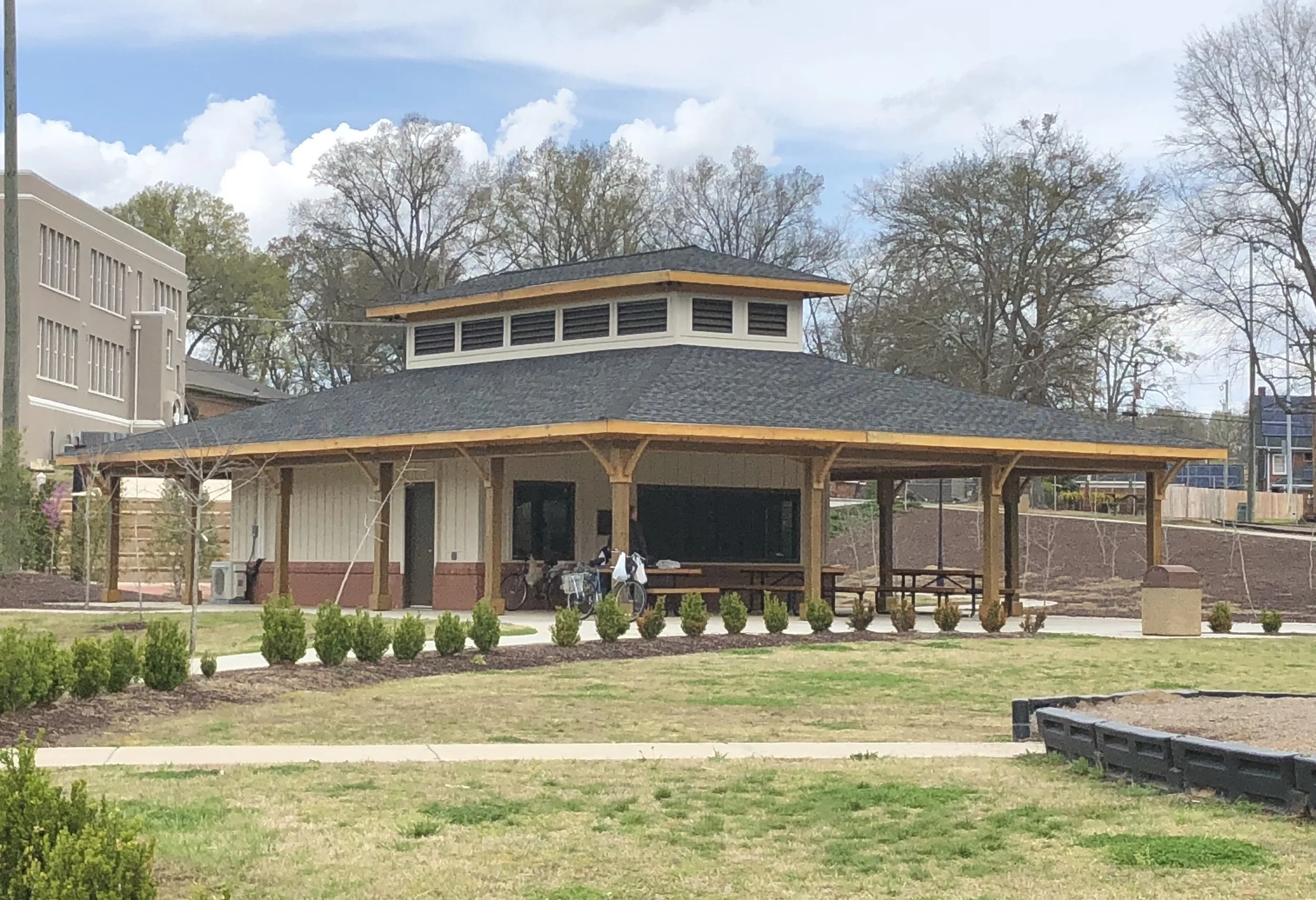 A park pavilion with a dark shingle roof and wooden beams, surrounded by a landscaped area with small bushes, bicycles, a bench, and a building with large windows behind it. Trees with fallen leaves are in the background under a partly cloudy sky.
