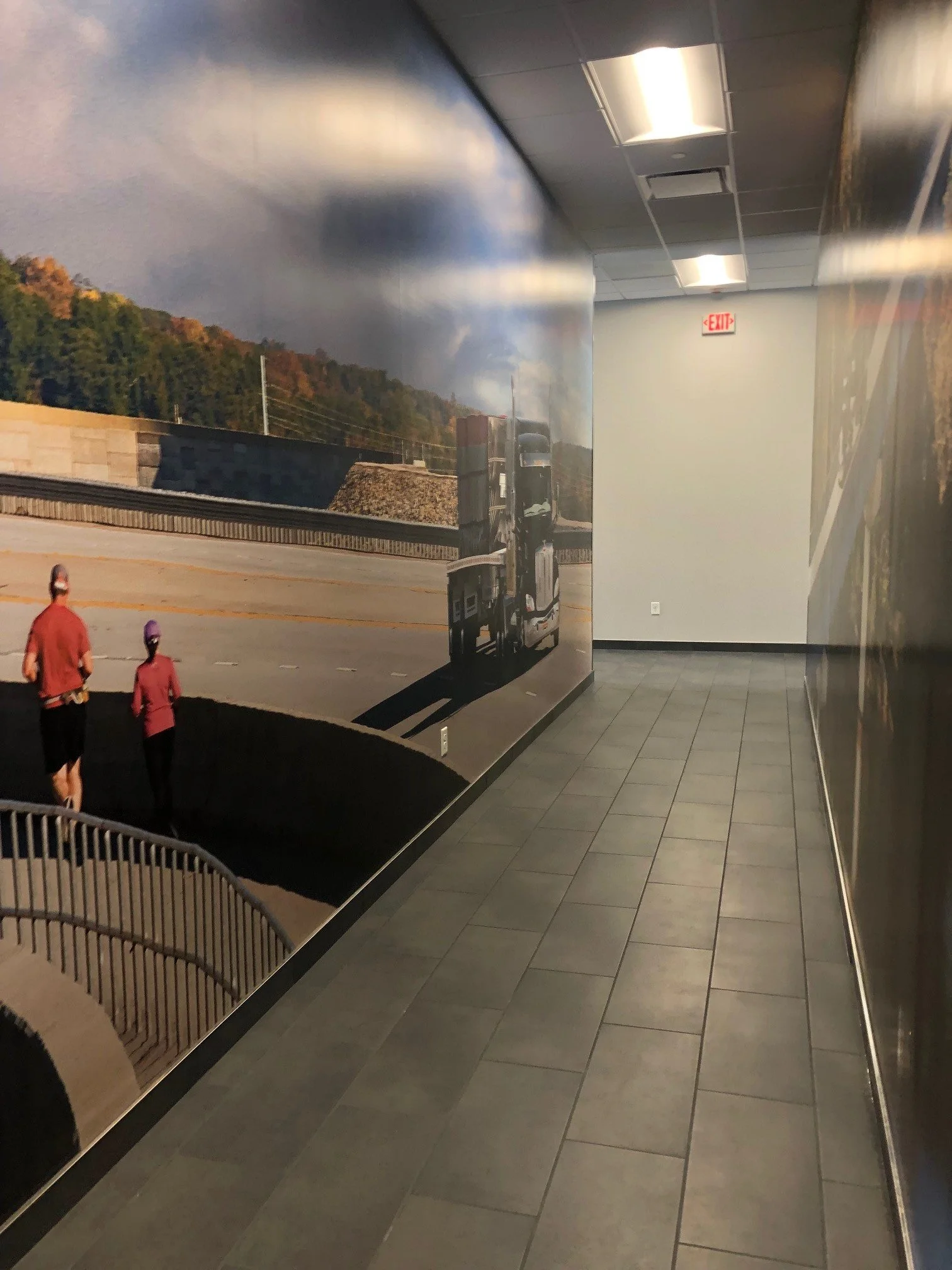 Indoor hallway with a collage mural of a stormy mountain landscape on the left wall, and gray tiled floor and ceiling with rectangular lights.