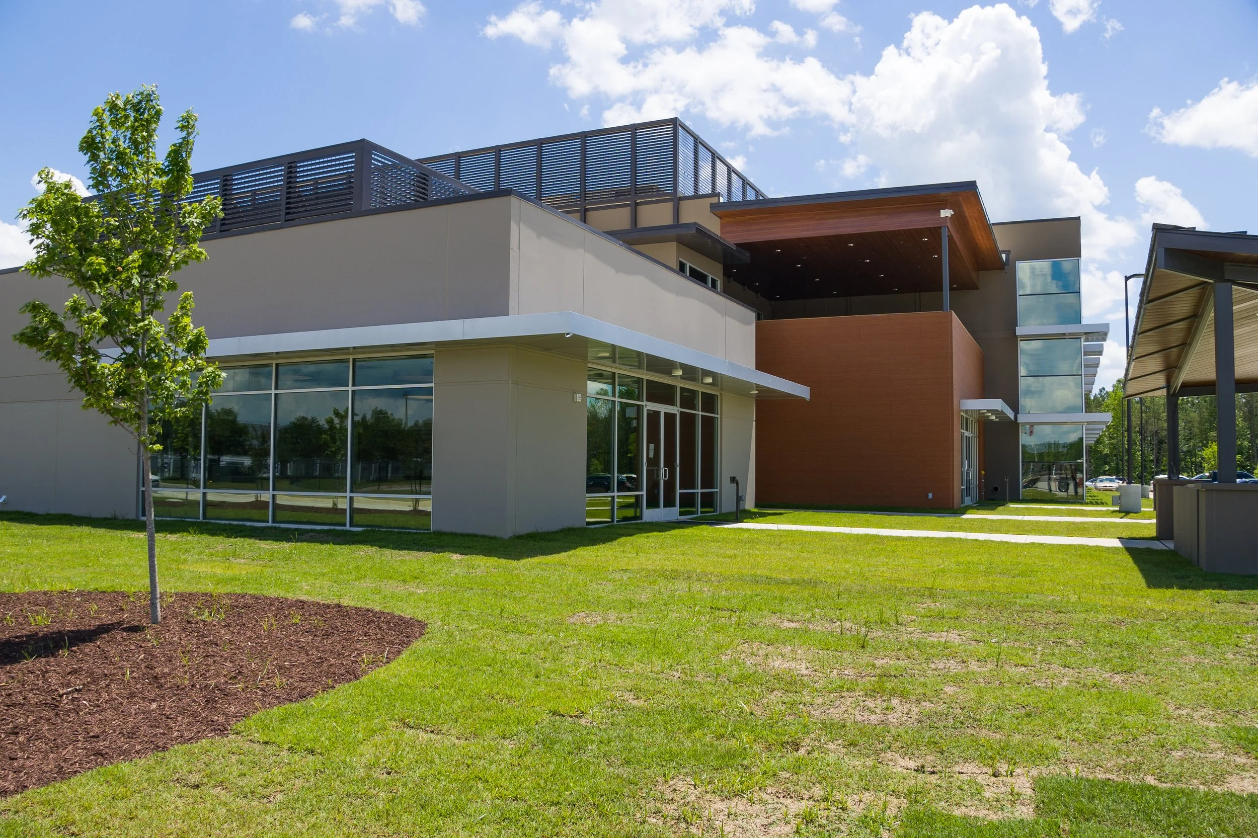 A modern multi-story building with large glass windows, a wooden overhang, and a spacious rooftop deck, set against a blue sky with scattered clouds and surrounded by a green lawn and small trees.
