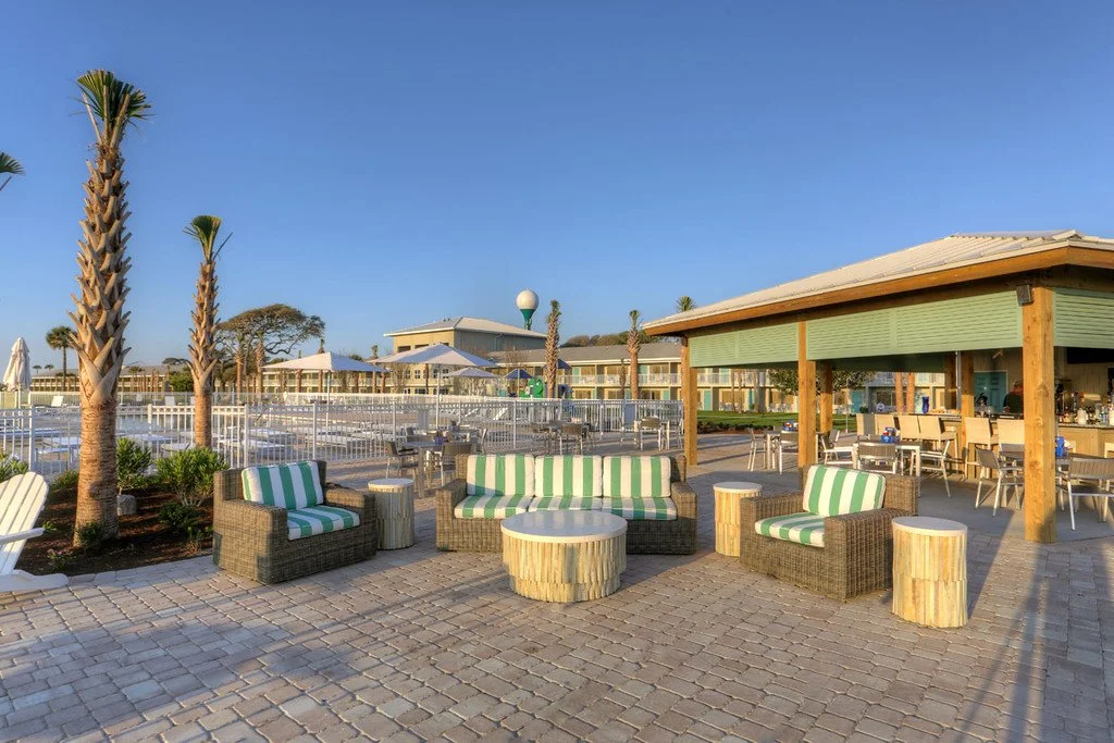 Outdoor lounge area with striped cushioned chairs and a round table, palm trees, and a poolside bar with tables and chairs, under a pavilion, with a clear blue sky.