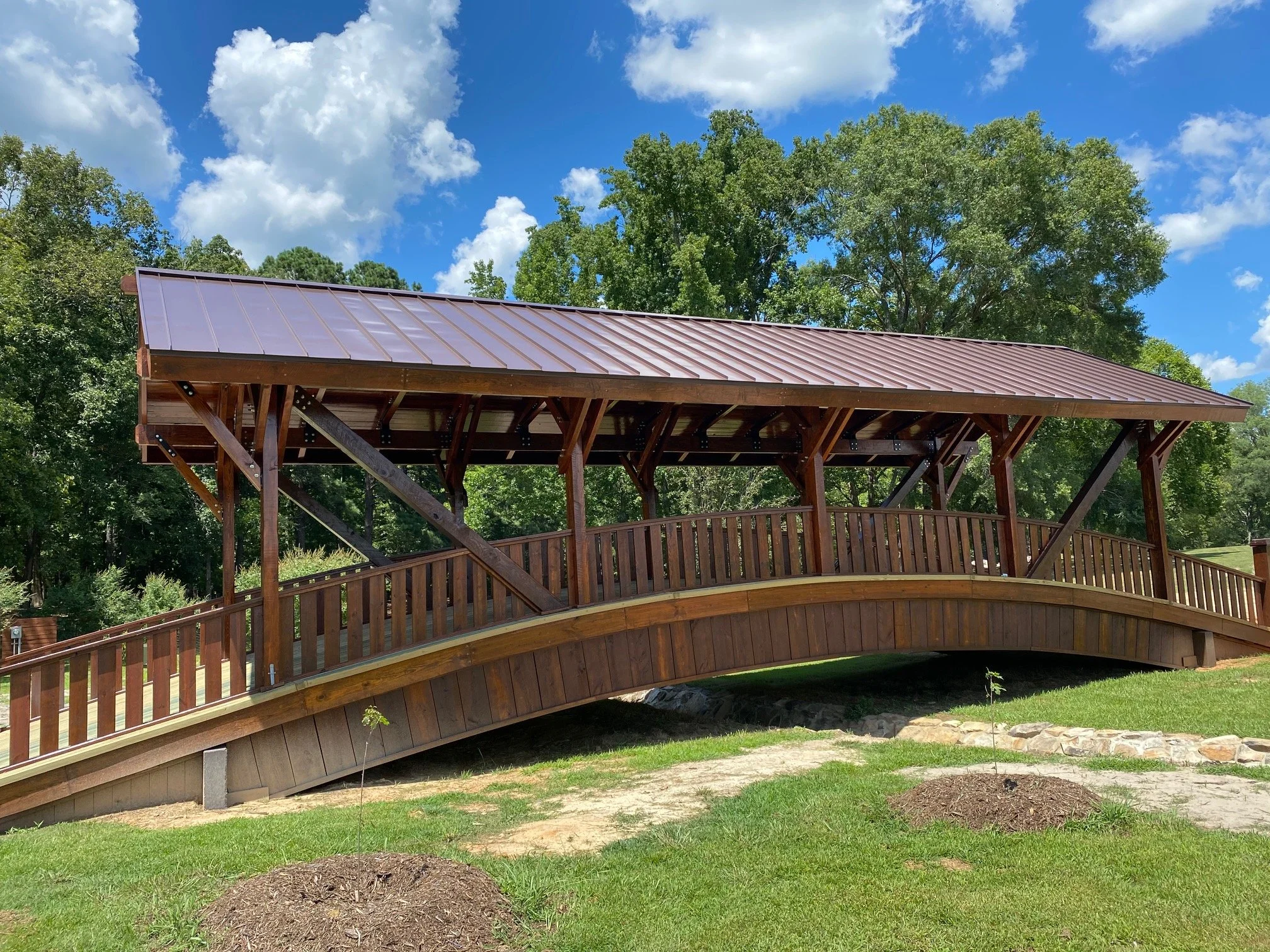 A wooden covered bridge with a metal roof, surrounded by green trees and grass, with a partly cloudy blue sky overhead.