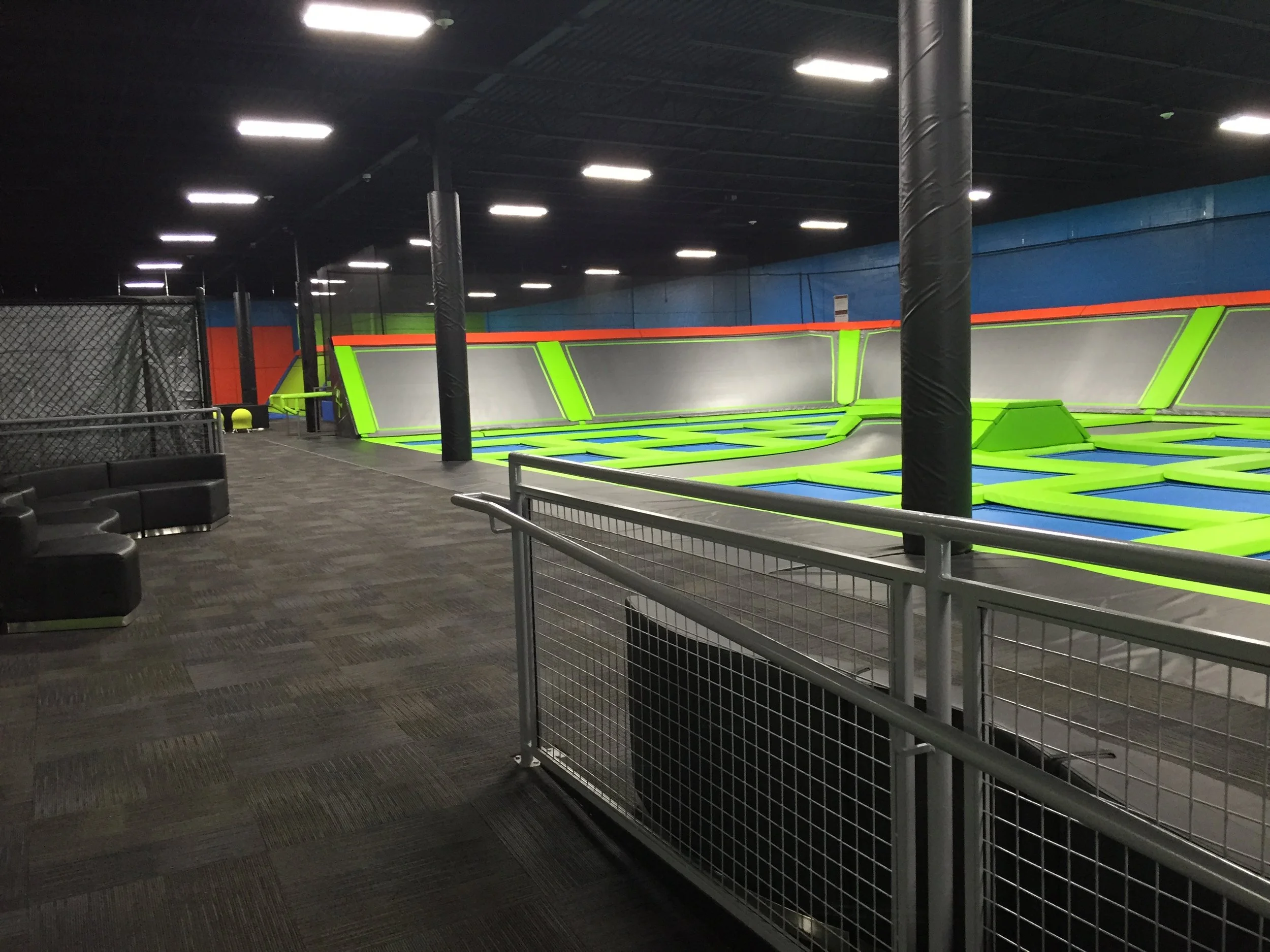 Indoor trampoline park with bright green and gray trampolines, black padded supports, and a black safety net. There are seating areas and a dark ceiling with bright lights.
