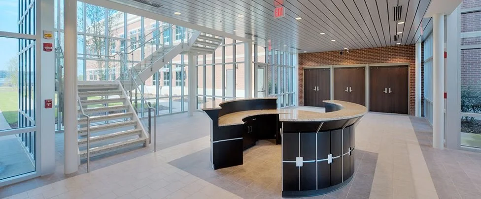 Interior of a modern building lobby with glass walls, a curved reception desk, brick wall, and a glass staircase.