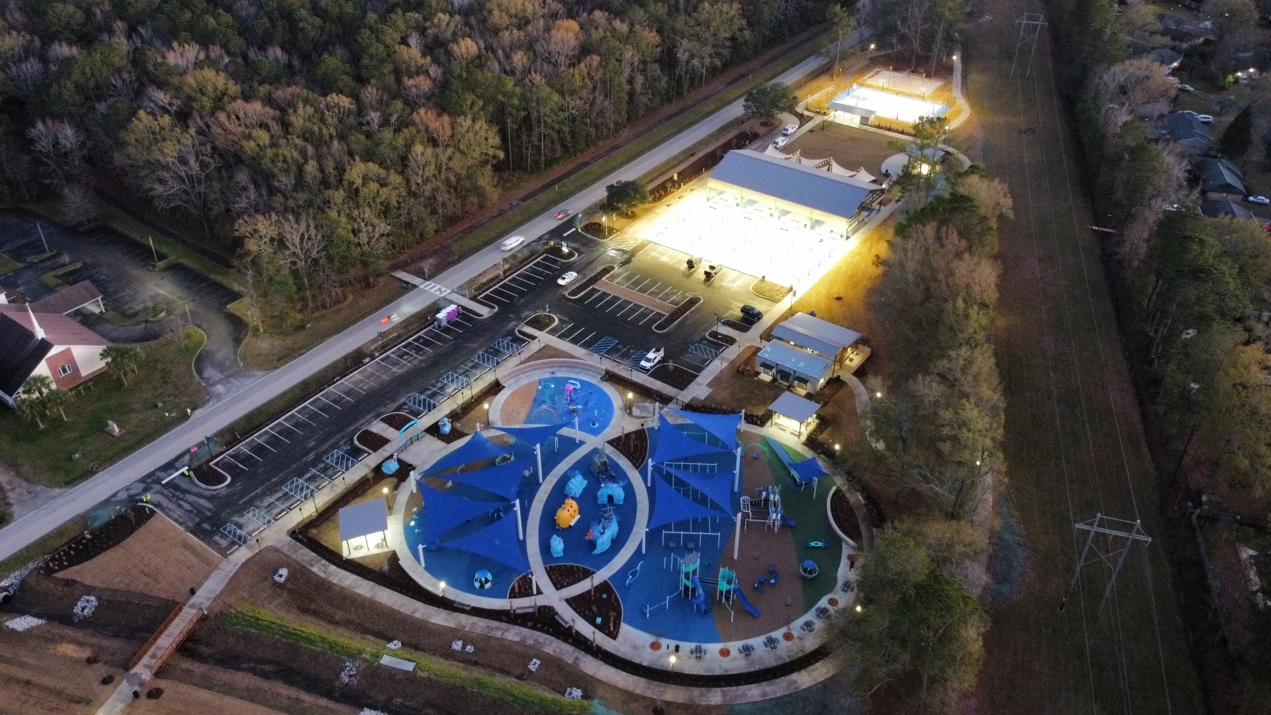 An aerial night view of a colorful playground with blue shade structures, a parking lot, a small building, and surrounding trees and residential houses.
