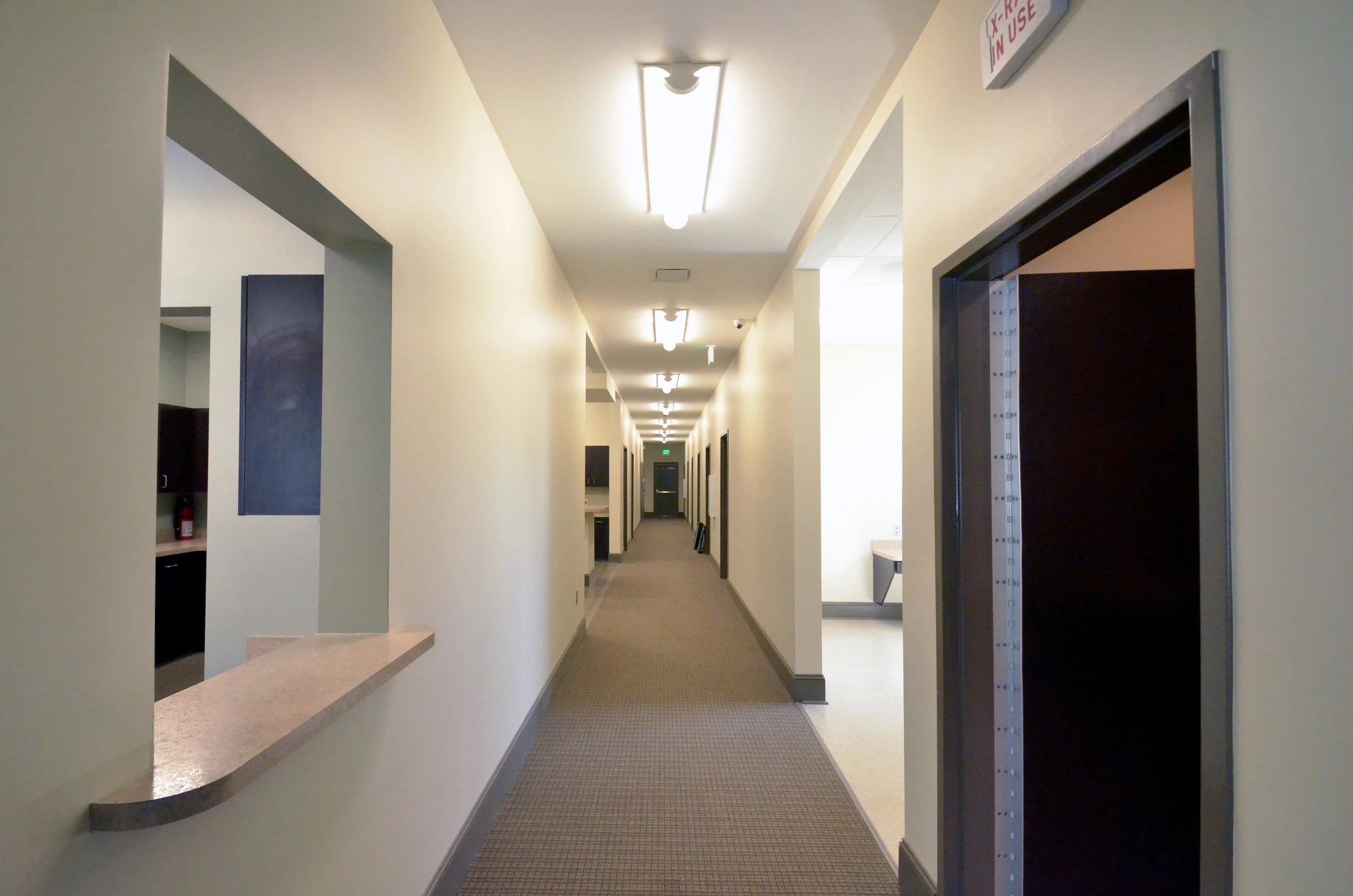 Long, well-lit hallway in a building, with doors on both sides, ceiling lights, and a fire extinguisher visible on the left.