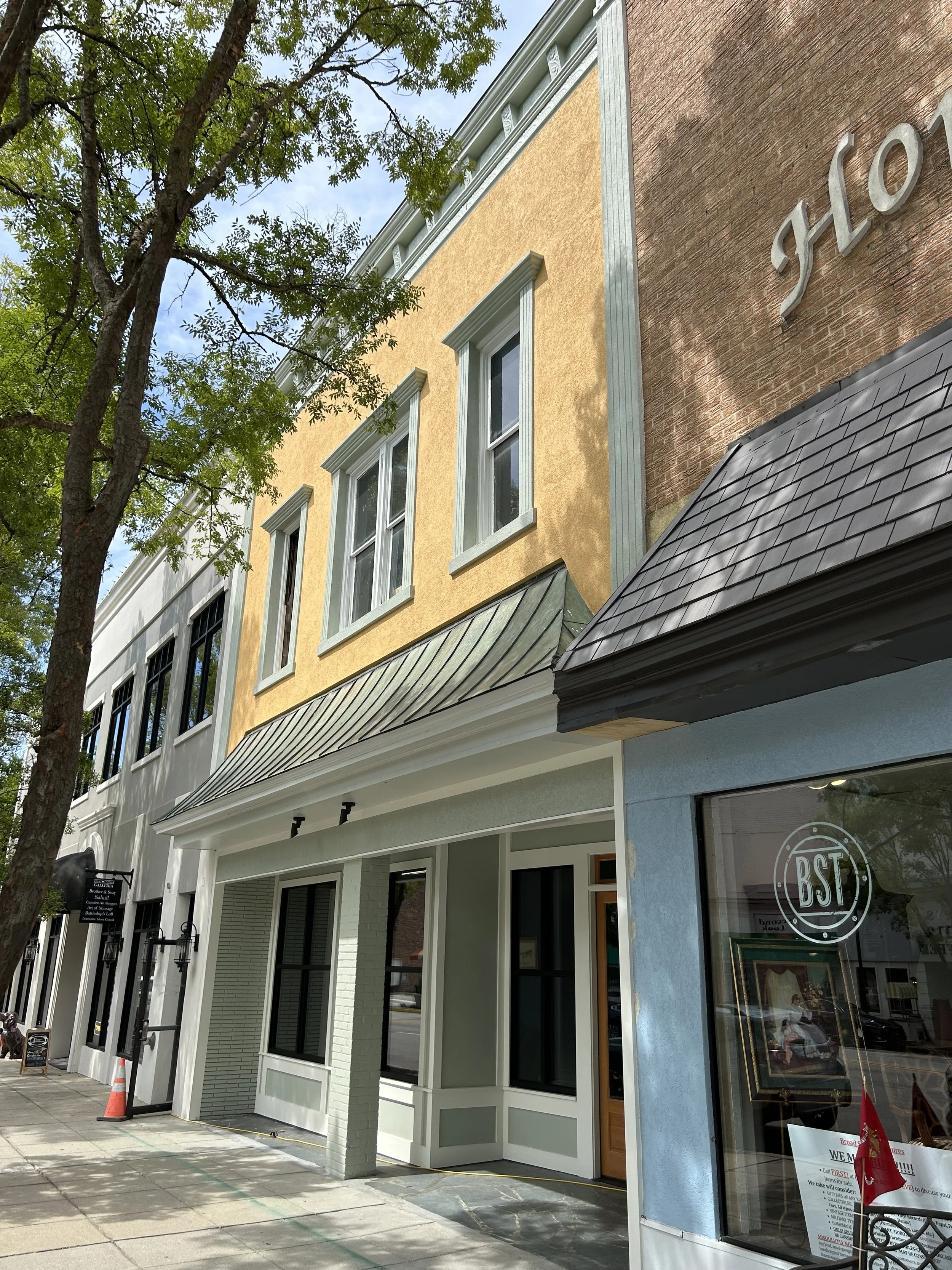 Street view of a row of storefronts with large window displays, a tree with green leaves, and a building with a yellow and a brick exterior. The storefront on the right has a sign with the logo 'BST' and a window with a sign and a small American flag.
