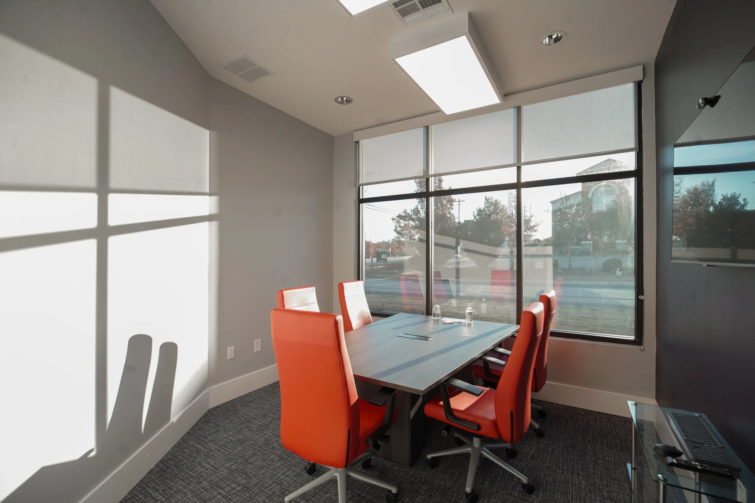 A modern conference room with a large table surrounded by orange chairs. Big windows let in natural light, casting shadows on the walls and table. Outside, trees and buildings are visible.