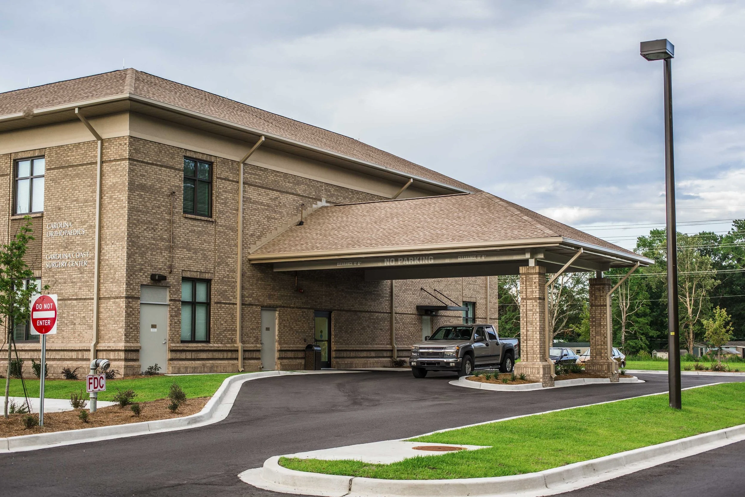The exterior of a medical building labeled "Carolina Orthopedics, Carolina Coast Surgery Center." There is a parking area with two vehicles, a black truck and a silver car. A tall streetlight and parking lot signs, including a "Do Not Enter" sign, ar