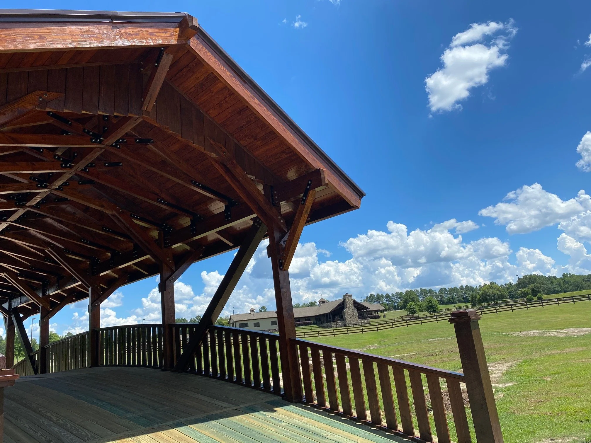 View of a covered wooden deck with a railing, overlooking a green field, houses, and a clear blue sky with some clouds.