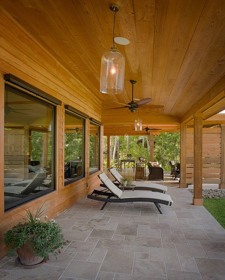 A covered outdoor patio with wood-paneled ceiling, large windows, two black and white lounge chairs with cushions, ceiling fans, hanging pendant lights, potted plant, and outdoor furniture in the background surrounded by trees.