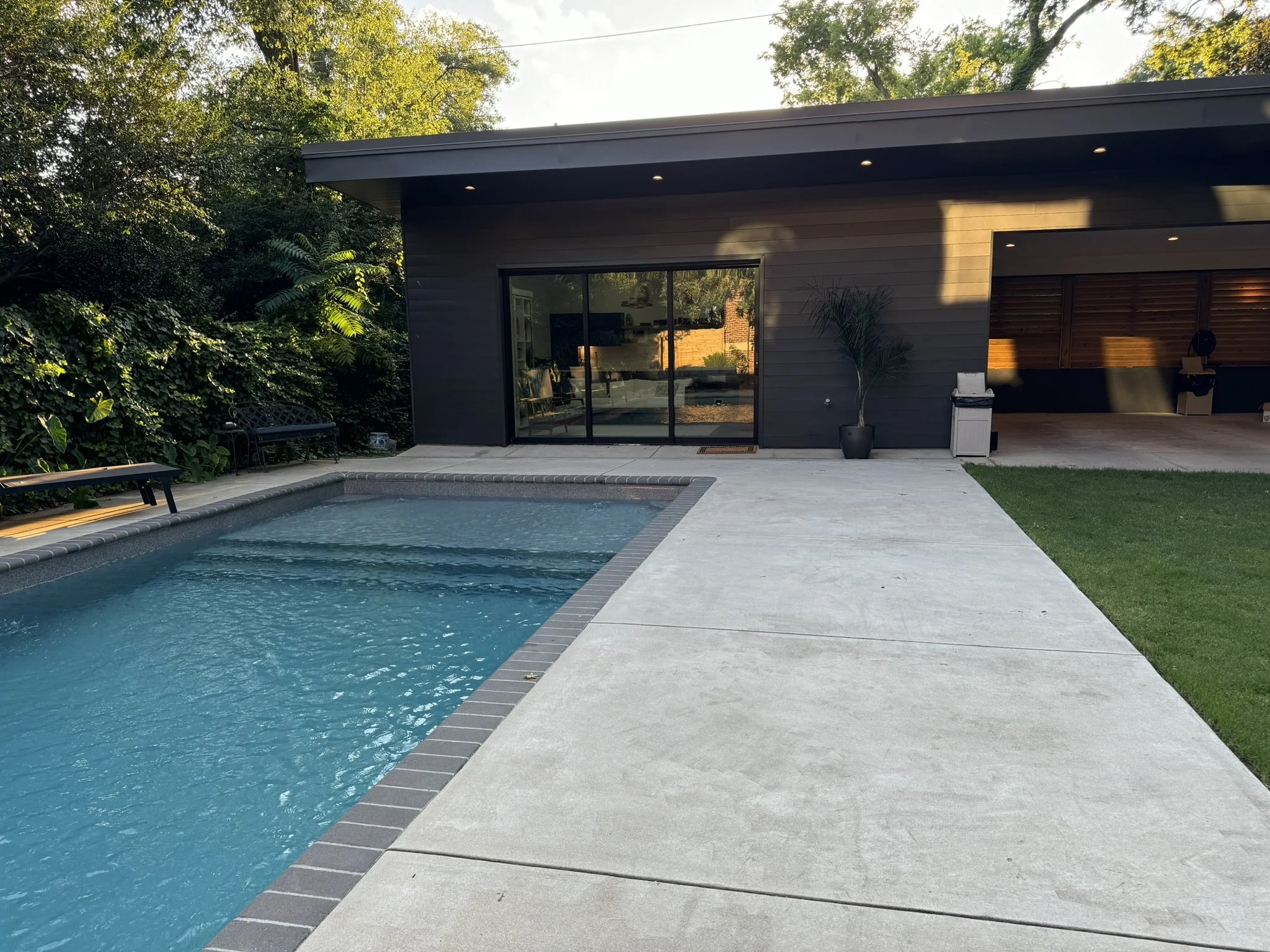 Backyard view with a swimming pool, concrete patio, modern house with sliding glass door, outdoor seating, and lush trees.
