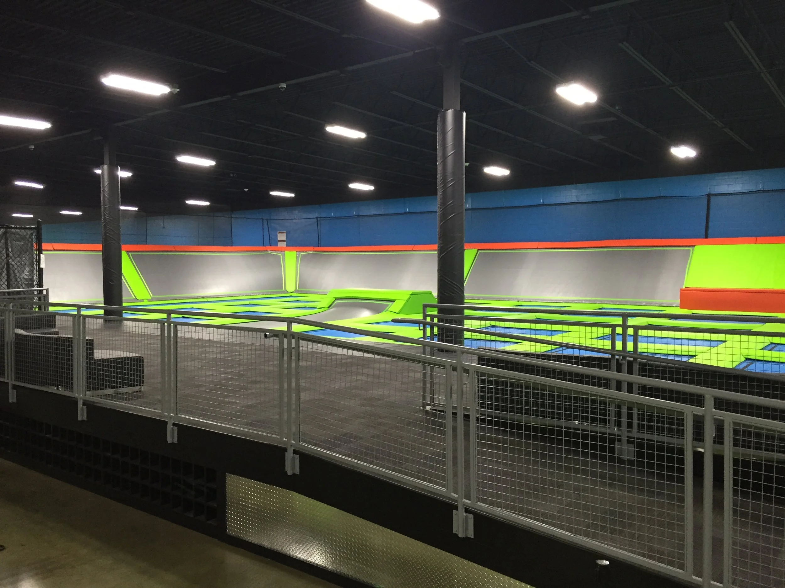 An indoor trampoline park with a large trampoline area bordered by a white safety fence, illuminated by overhead fluorescent lights, with blue and green padded walls and a black ceiling.