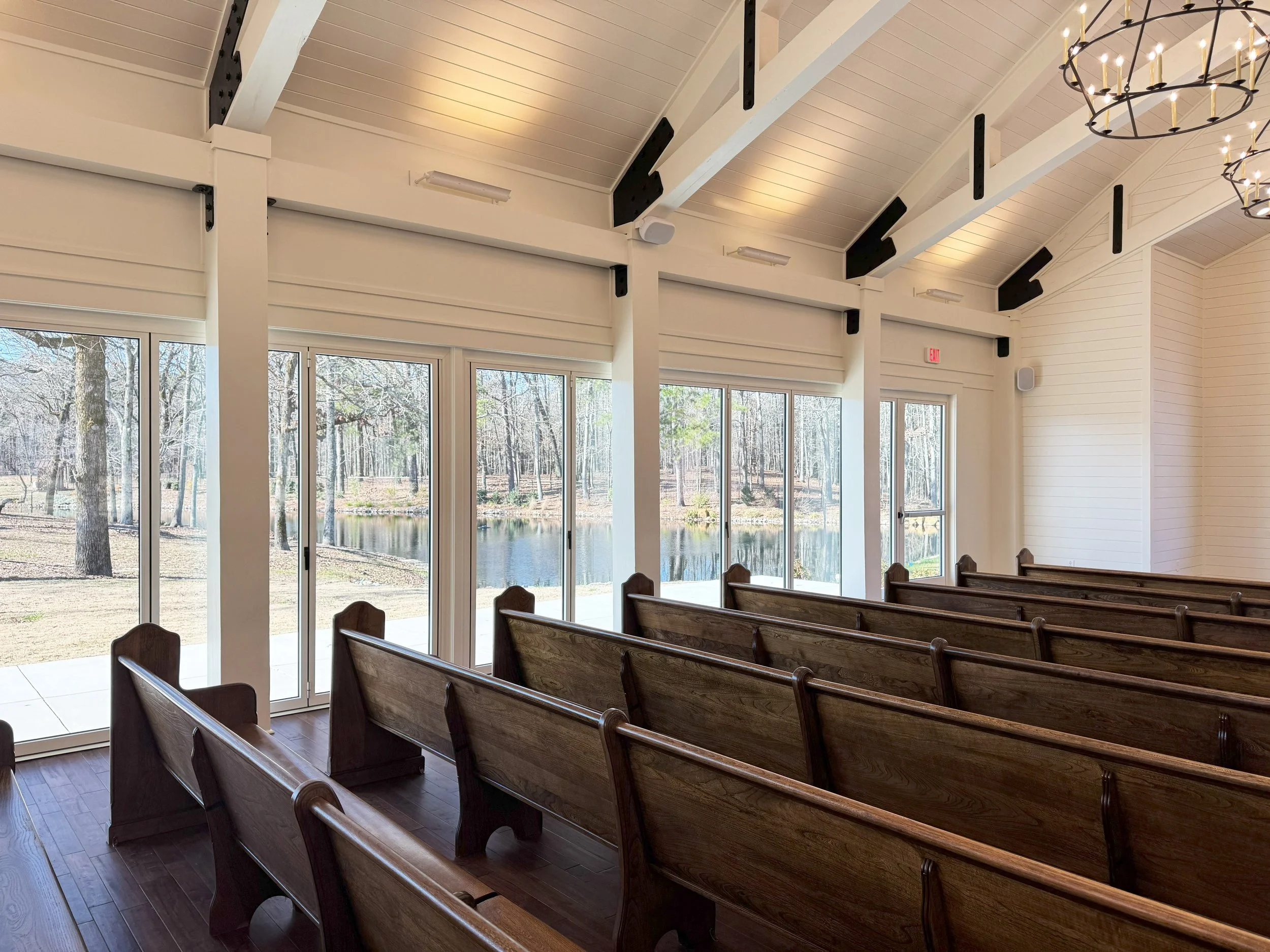 Interior of a church with wooden pews, large glass windows, white paneled walls, and a high white ceiling with beams and chandeliers, overlooking an outdoor wooded area.