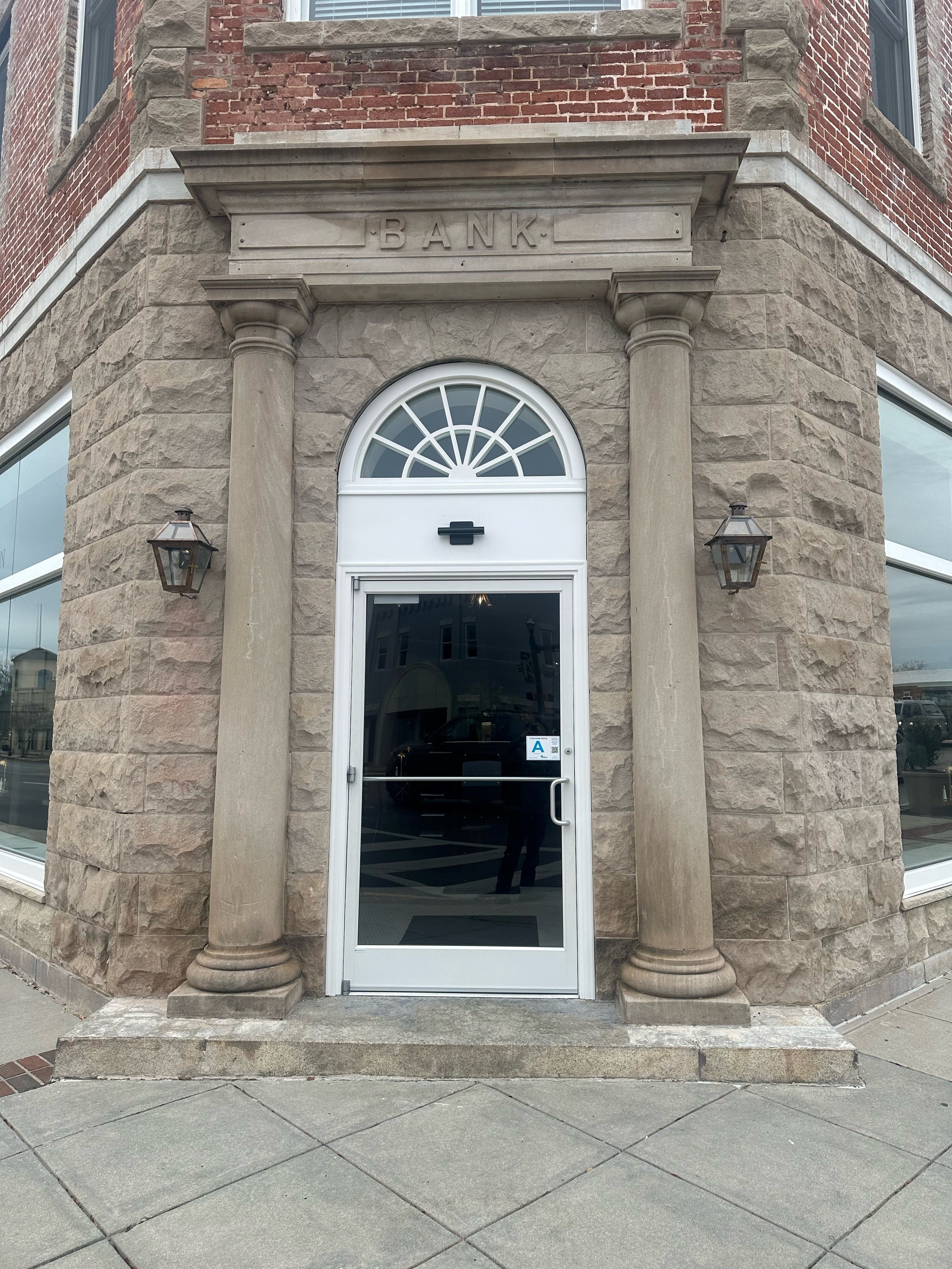 Front entrance of a building with a white door, surrounding stone columns, and a plaque above the door area that says 'BANK'. Two wall-mounted lantern-style lights flank the door, and there are brick and stone masonry details on the building's exteri