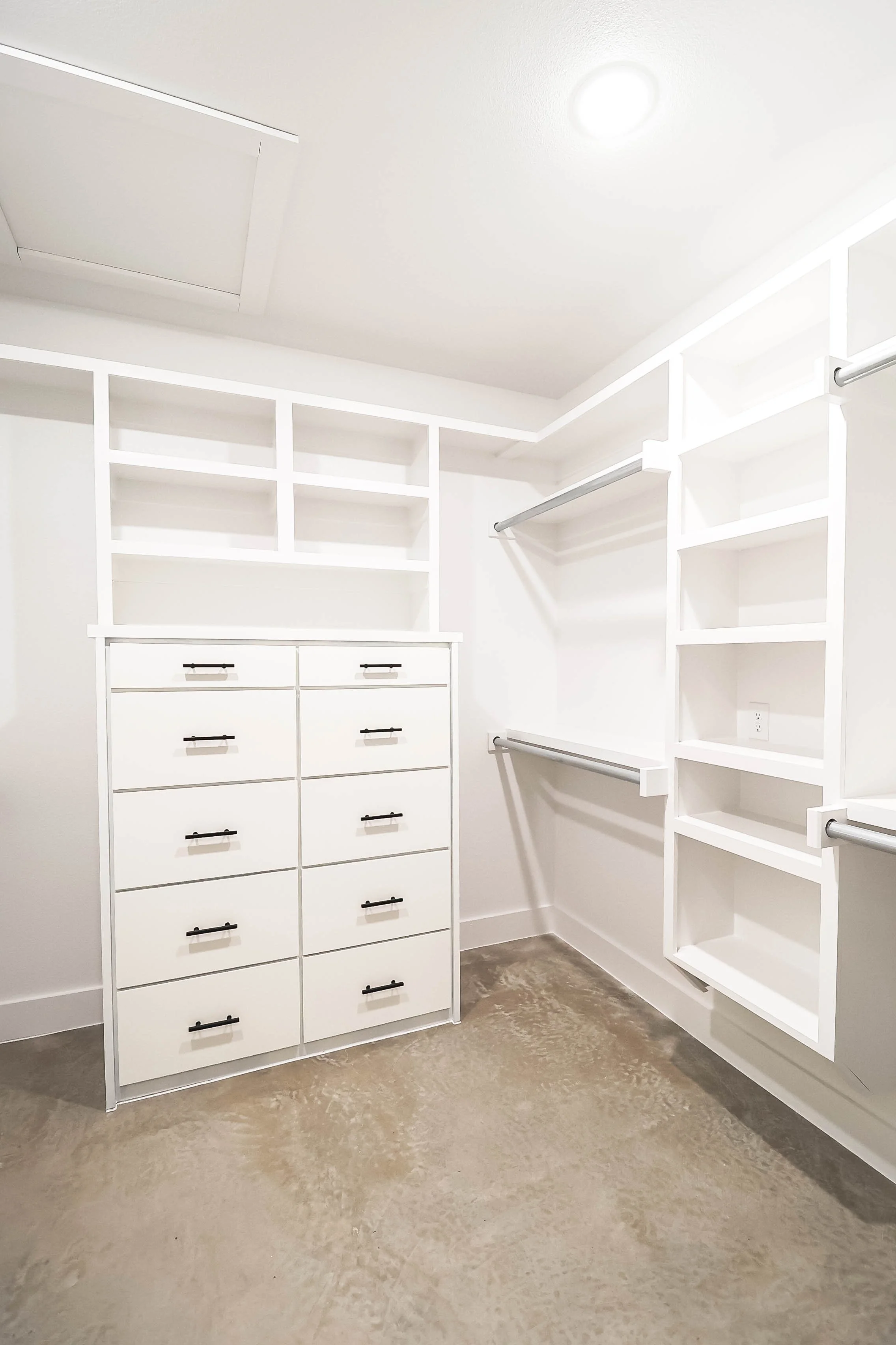 Empty walk-in closet with white cabinetry, open shelves, drawers, and metal hanging rods, beige concrete floor, and white walls.