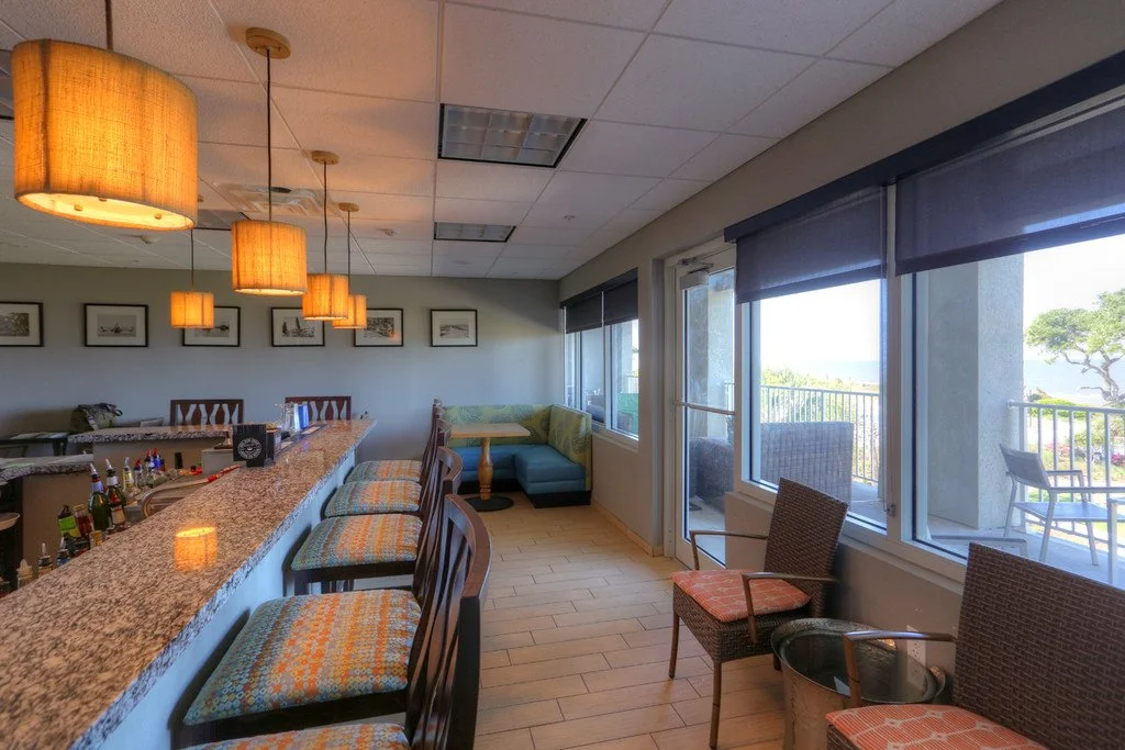 Indoor lounge area with bar counter, chairs, and large windows looking out to a balcony and greenery.