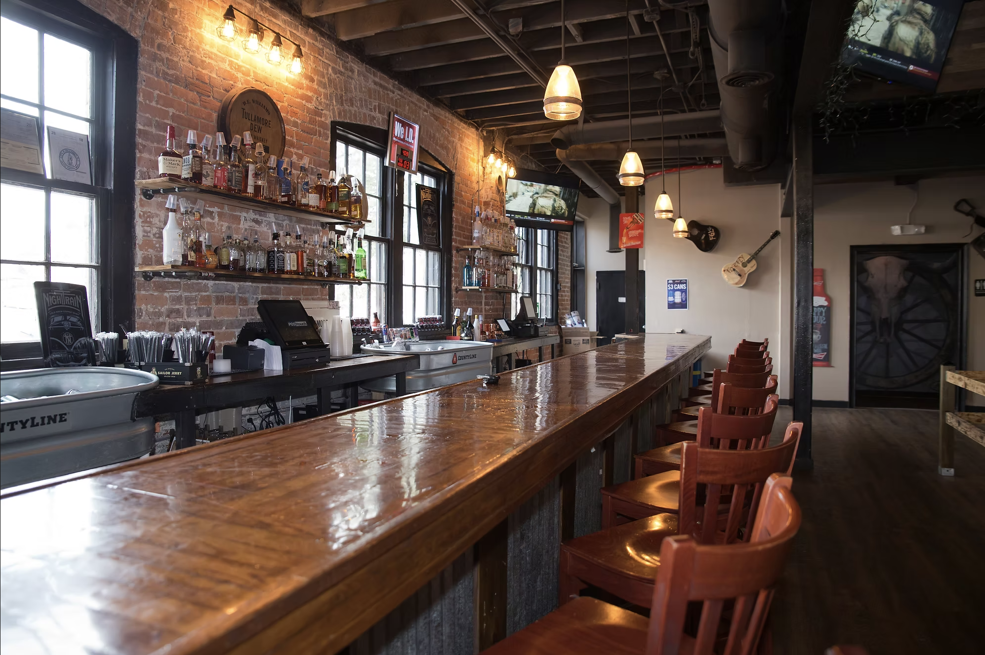 Interior of a bar with a long wooden counter, brick walls, and several bottles of alcohol on shelves. There are hanging pendant lights, a television, and a guitar mounted on the wall.