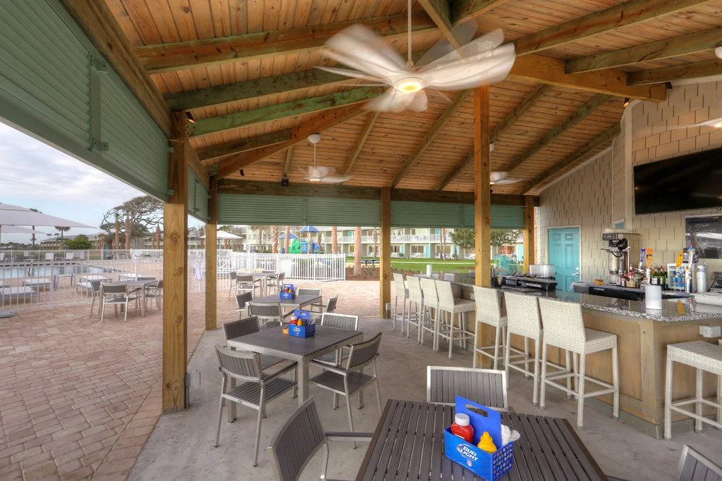 An outdoor seating area at a bar or restaurant with a wooden ceiling, ceiling fans, bar counter, and several tables with condiments, overlooking a pool or recreation area with umbrellas and lounge chairs.