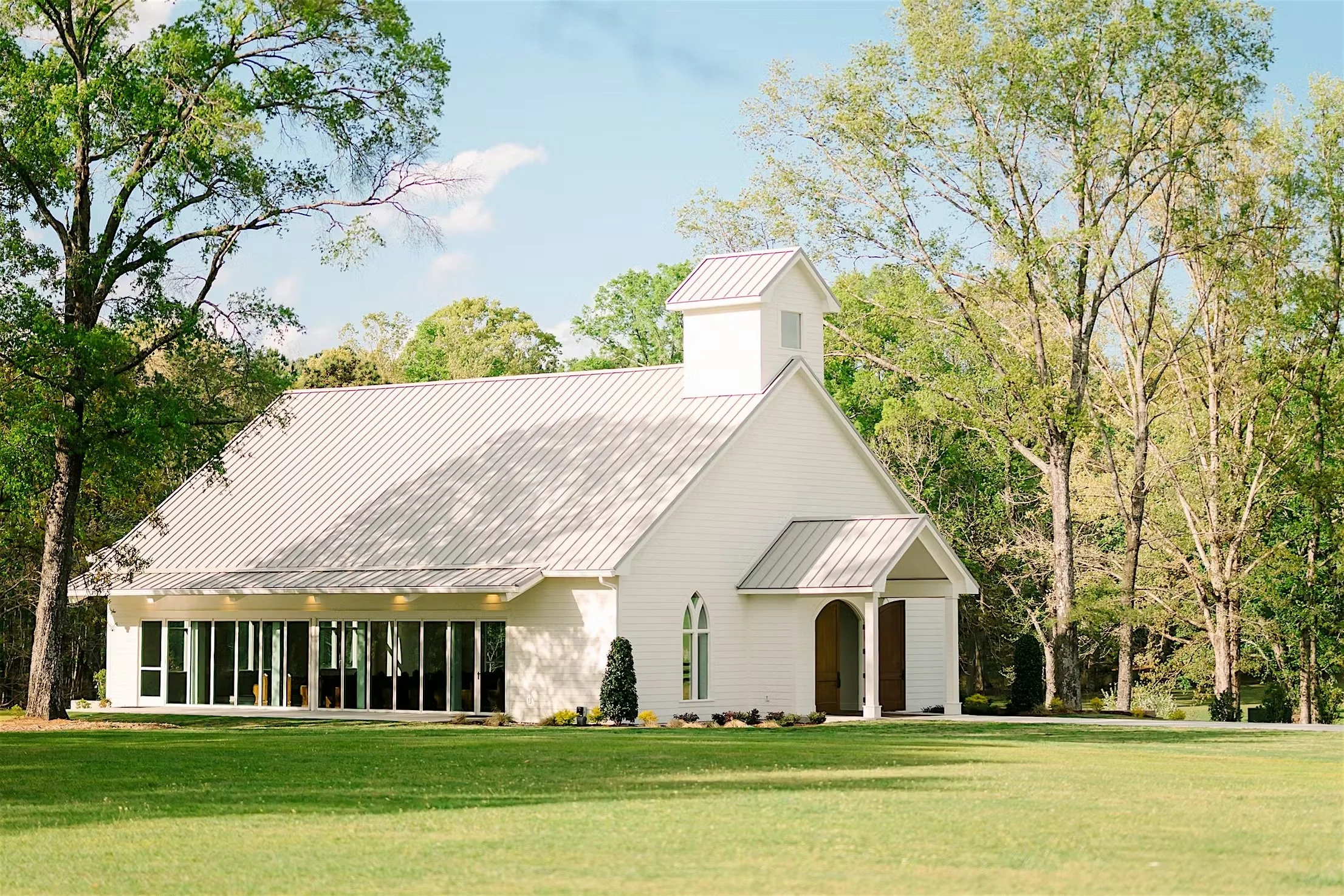 White church with a steeple, surrounded by tall trees with green leaves, a green lawn, and a partly cloudy blue sky.